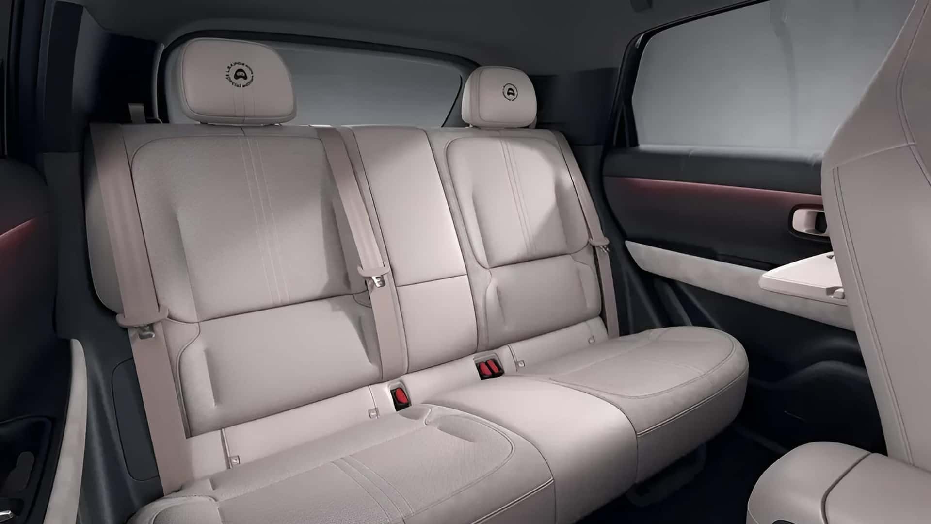 Interior de carro com bancos traseiros em couro bege claro, cintos de seguran&ccedil;a vis&iacute;veis e encostos de cabe&ccedil;a com um logotipo circular. A porta lateral tem detalhes em preto e uma faixa iluminada em vermelho, com uma pequena mesa dobr&aacute;vel no encosto do banco dianteiro