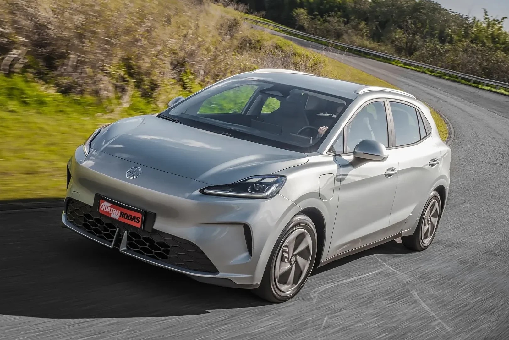 Carro elétrico prateado, modelo SUV compacto, em movimento numa estrada sinuosa com vegetação verde e céu azul ao fundo