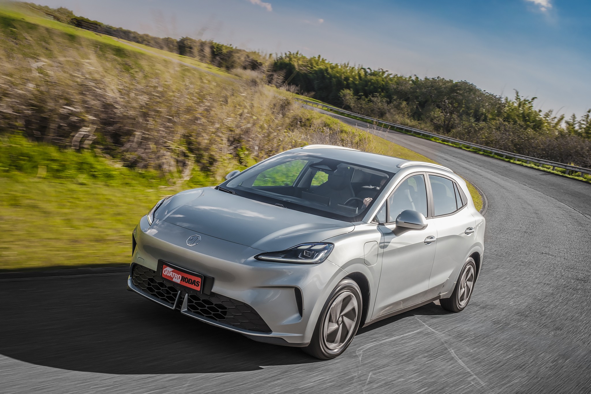 Carro el&eacute;trico prateado em movimento numa estrada sinuosa, com vegeta&ccedil;&atilde;o verde e c&eacute;u azul ao fundo