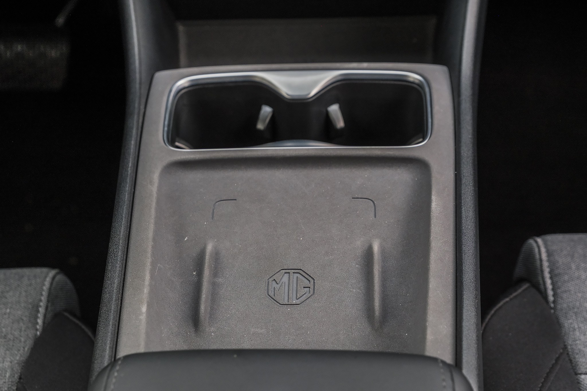 Console central de carro cinza escuro com dois porta-copos cromados e um compartimento de carregamento sem fio com o logotipo MG em relevo