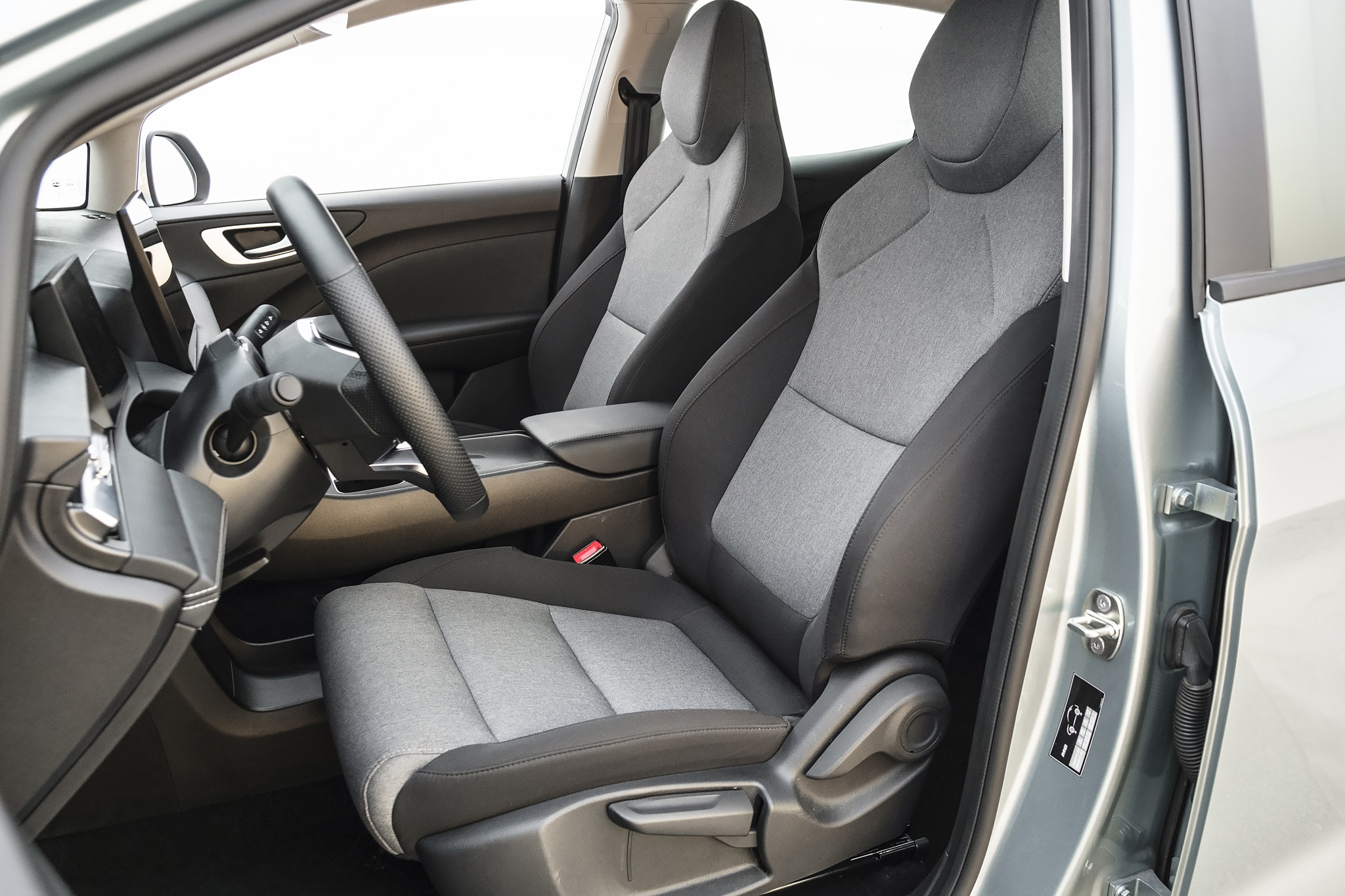 Interior de um carro com bancos dianteiros cinza claro e escuro, volante preto texturizado e painel escuro, visto da porta aberta do lado do passageiro