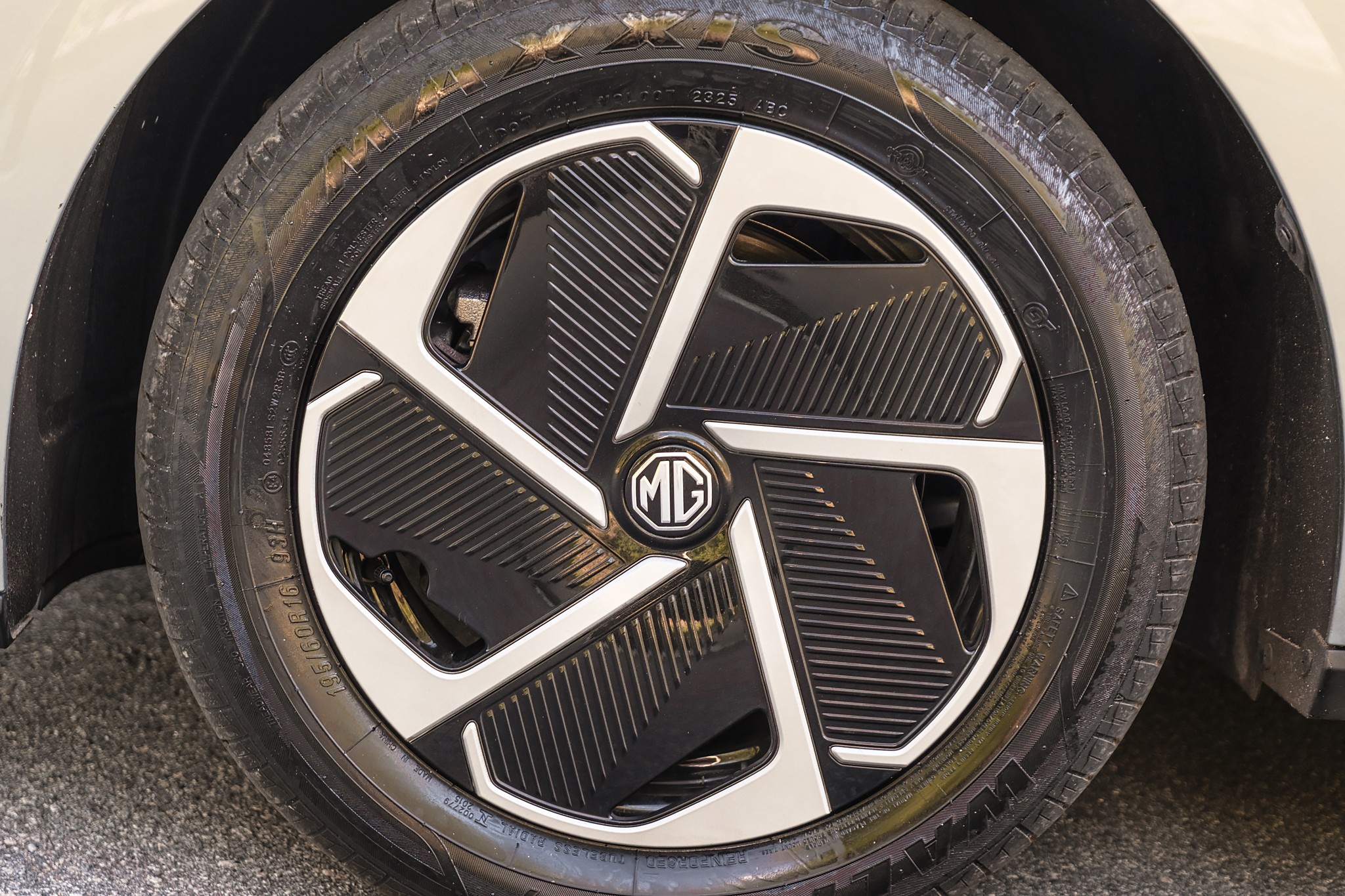Pneu de carro com calota preta e detalhes prateados em formato de h&eacute;lice, com o logotipo MG ao centro, sobre asfalto cinza