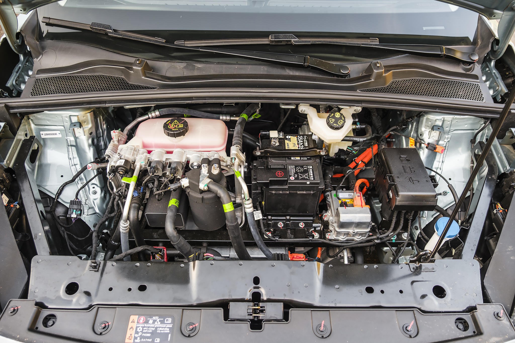 Compartimento do motor de um carro el&eacute;trico, com bateria preta, reservat&oacute;rios de fluidos rosa e branco, e cabos laranja e pretos vis&iacute;veis