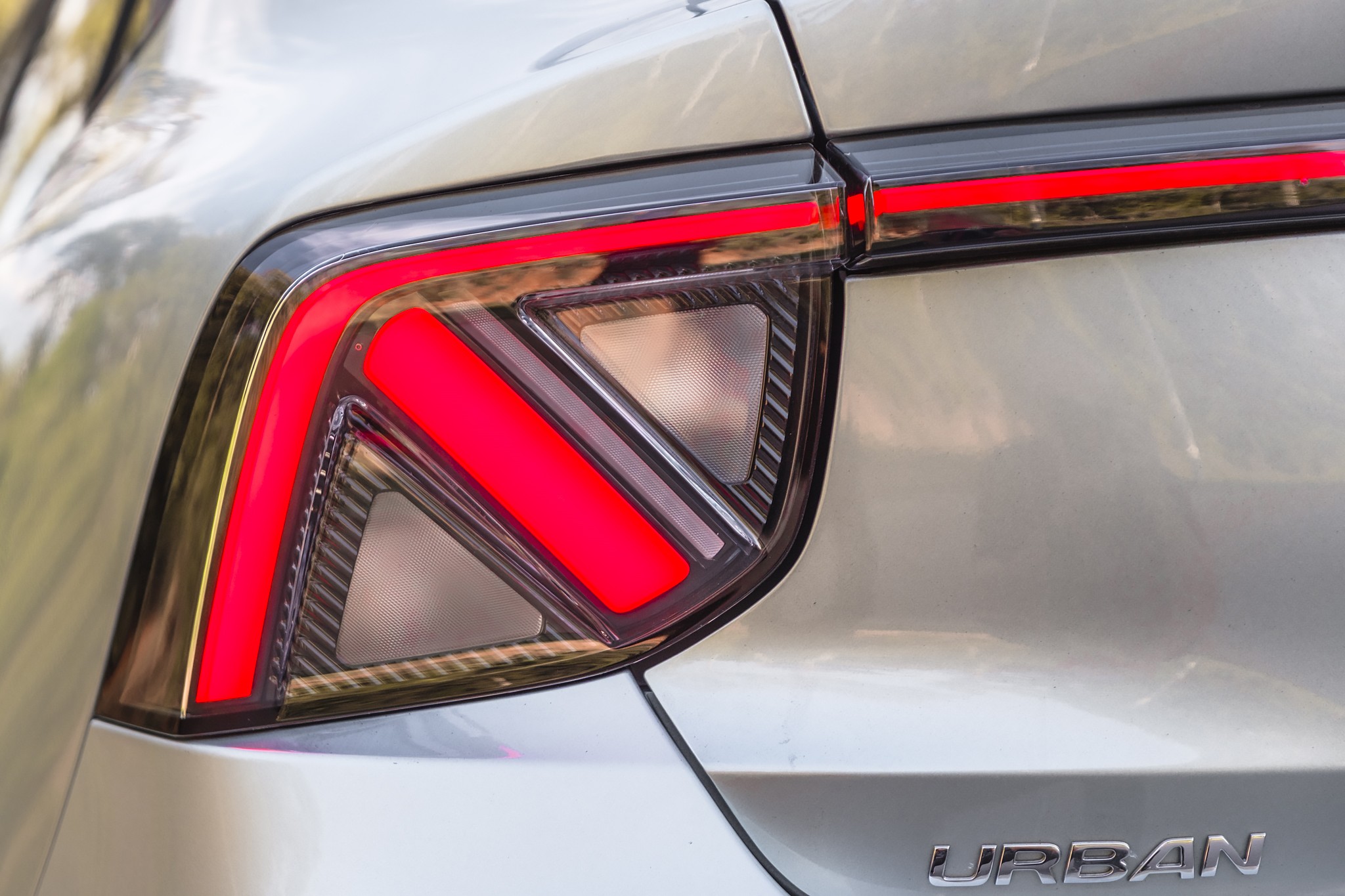 Lanterna traseira direita de um carro cinza-claro, com luzes vermelhas acesas em formato de L e uma barra horizontal. O nome URBAN est&aacute; em relevo prateado na parte inferior direita