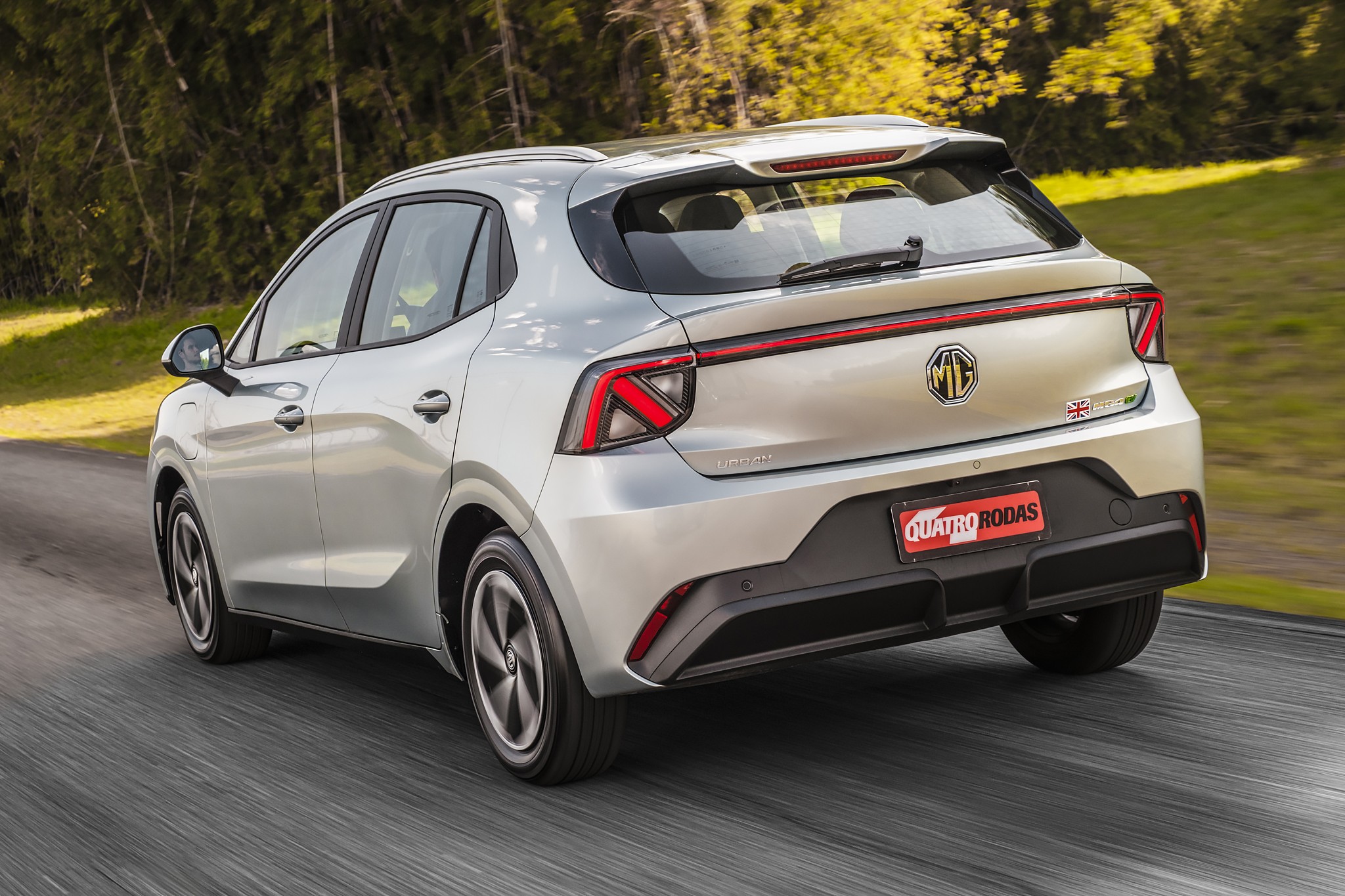 Carro prata MG, modelo SUV compacto, visto de traseira e lateral, em movimento numa estrada asfaltada. O ve&iacute;culo tem lanternas traseiras em formato de Z e para-choque preto. Ao fundo, &aacute;rvores e vegeta&ccedil;&atilde;o verde emba&ccedil;adas pelo movimento