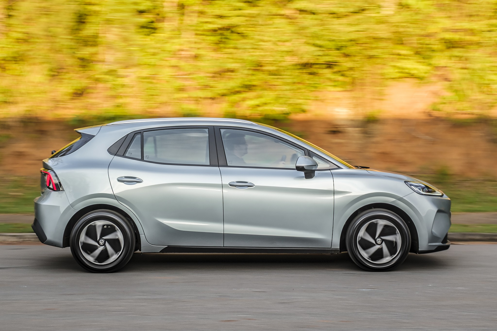 Carro hatch prateado em movimento, visto de lado, com fundo de vegeta&ccedil;&atilde;o borrado pelo movimento. As rodas t&ecirc;m design de h&eacute;lice preta e prata.
