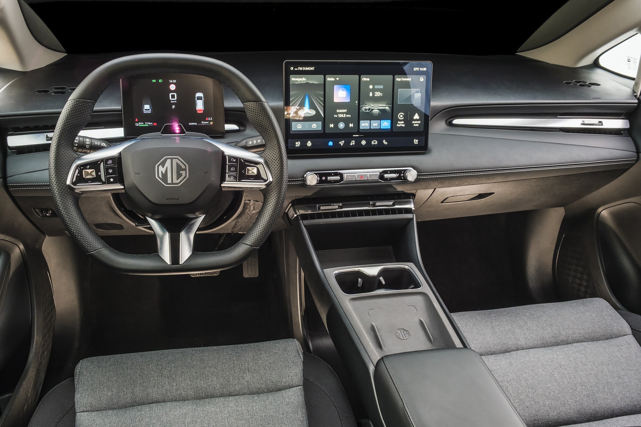 Interior moderno de um carro, com volante multifuncional MG, painel digital e tela multim&iacute;dia central exibindo aplicativos. Bancos cinza claro e console central com porta-copos.