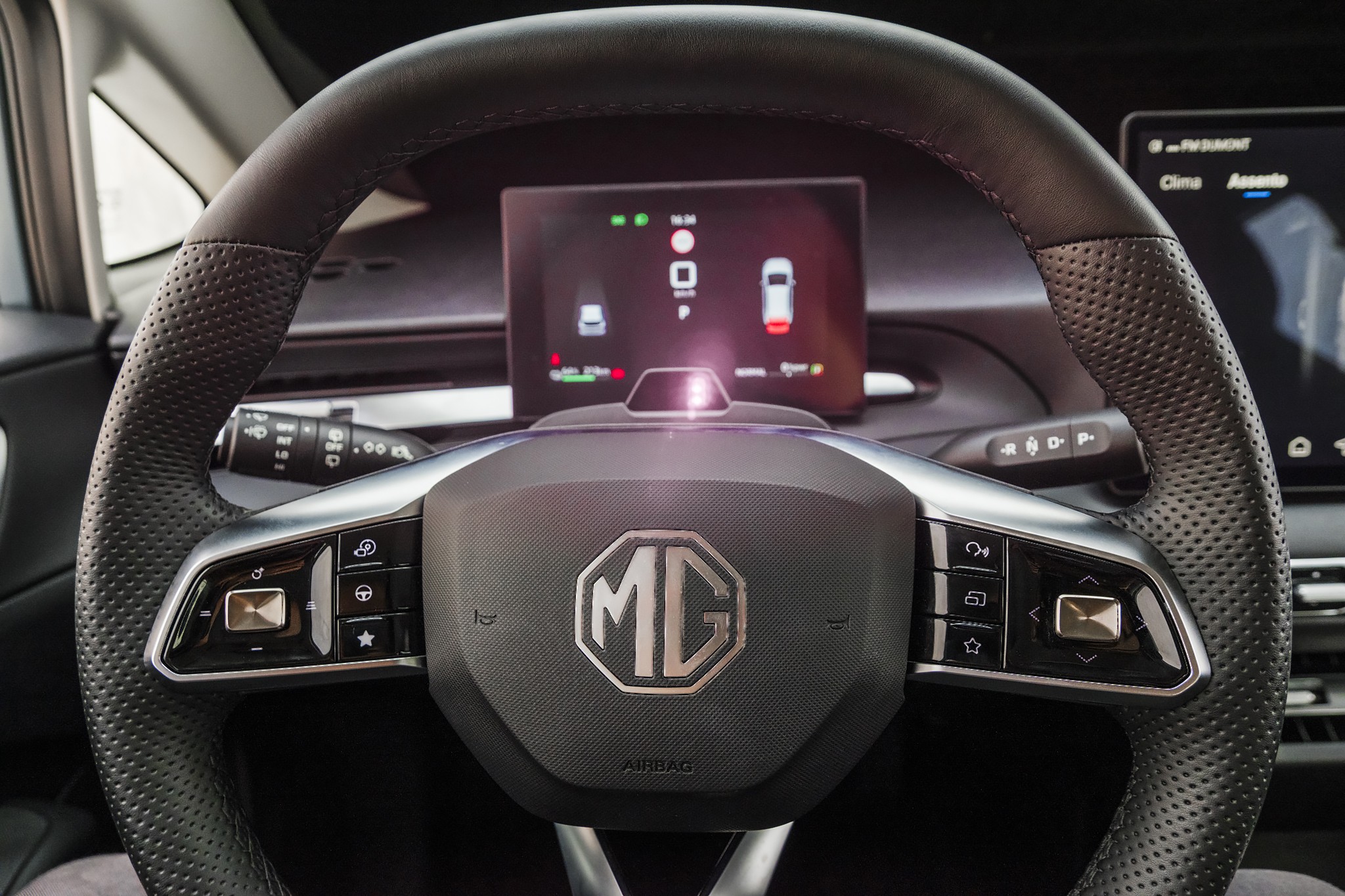 Volante de carro moderno em couro preto perfurado, com bot&otilde;es de controle prateados e o logotipo MG no centro. Ao fundo, um painel digital exibe informa&ccedil;&otilde;es de assist&ecirc;ncia ao motorista, como dist&acirc;ncia de seguran&ccedil;a e alerta de colis&atilde;o, com &iacute;cones de ve&iacute;culos e luzes indicadoras