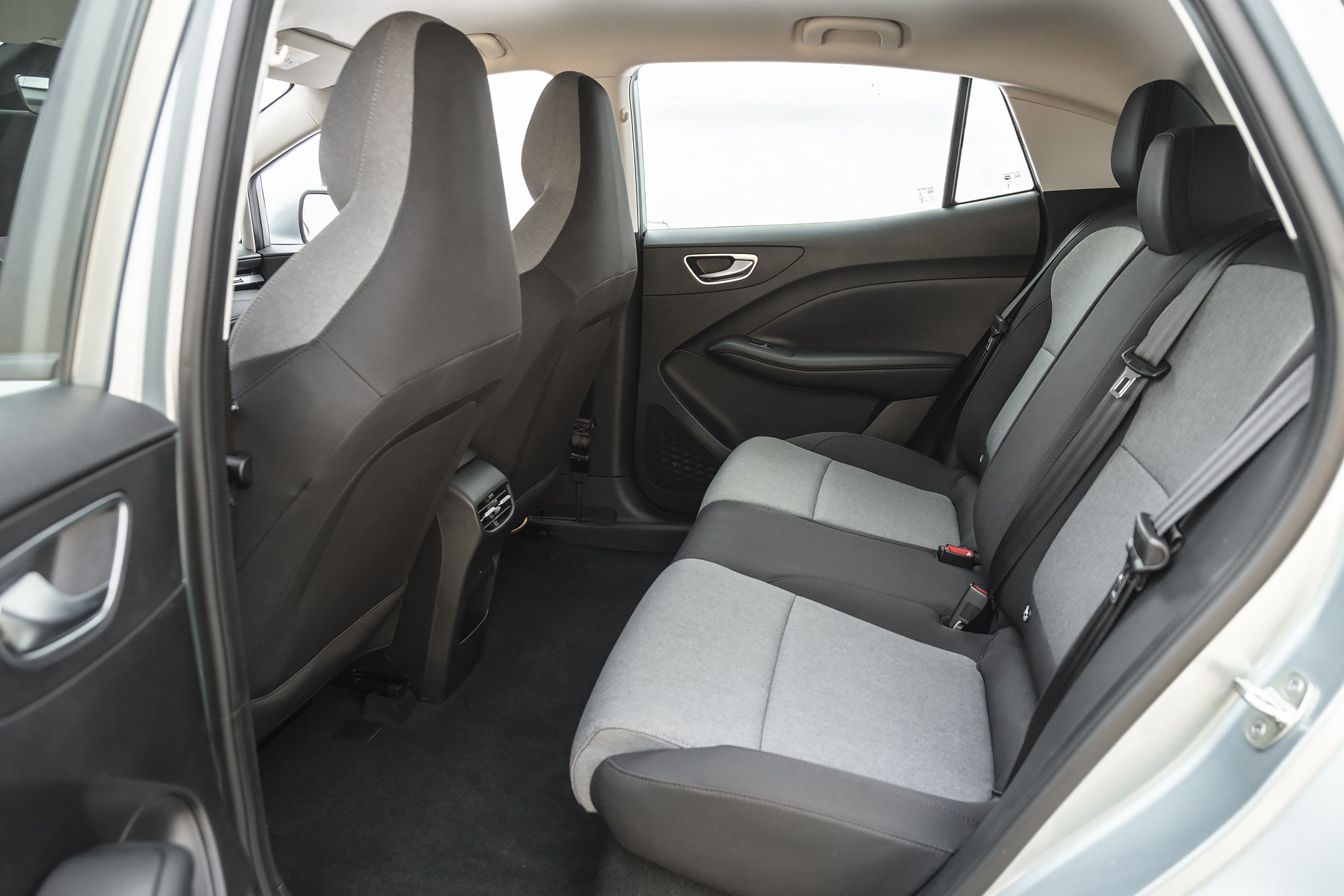Interior de carro com bancos traseiros em tecido cinza claro e escuro, cintos de seguran&ccedil;a vis&iacute;veis e encostos de cabe&ccedil;a pretos. As portas e o piso s&atilde;o escuros, com detalhes cromados nas ma&ccedil;anetas. A janela traseira mostra um c&eacute;u claro