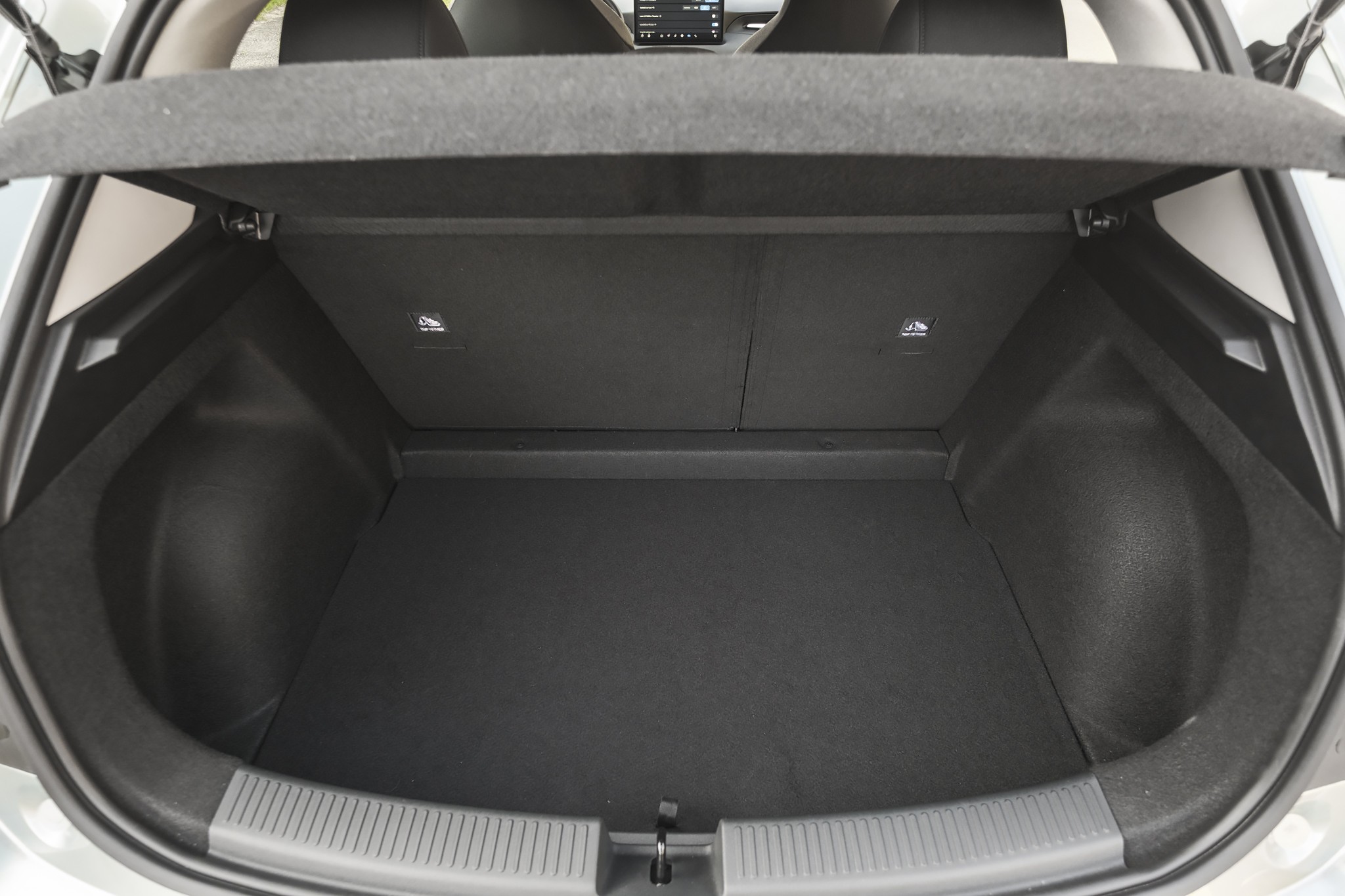 Porta-malas de um carro hatch, visto de cima, com o compartimento vazio e forrado em carpete cinza escuro. O tamp&atilde;o do porta-malas est&aacute; levantado, revelando o interior do ve&iacute;culo e parte dos bancos traseiros