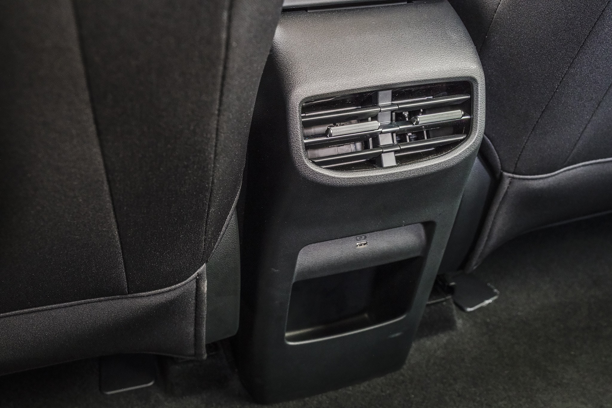Sa&iacute;da de ar condicionado traseira em um carro, com uma porta USB e um pequeno porta-objetos abaixo, entre dois bancos pretos