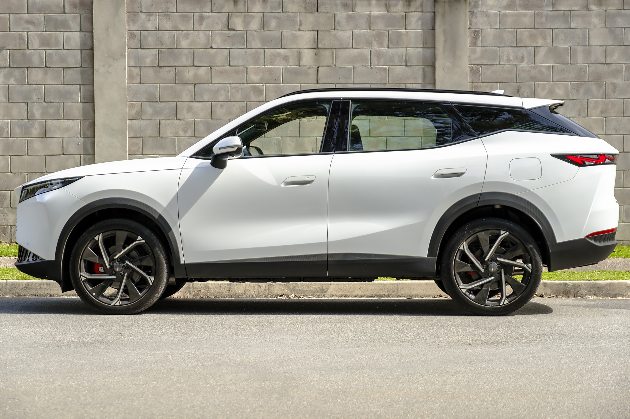 SUV branco moderno, com teto preto e rodas esportivas escuras, estacionado de perfil em uma rua asfaltada, com um muro de tijolos cinzas ao fundo
