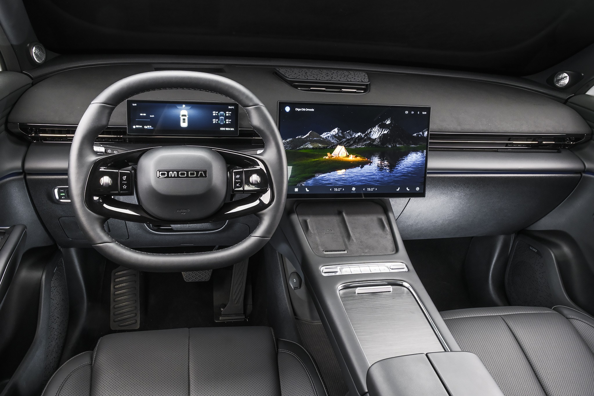 Interior de um carro moderno com volante preto multifuncional e logo OMODA. Dois displays digitais: um menor atr&aacute;s do volante mostrando informa&ccedil;&otilde;es do ve&iacute;culo, e outro maior &agrave; direita exibindo uma paisagem noturna com montanhas, lago e uma barraca iluminada. O console central possui acabamento escuro e detalhes prateados, com bancos de couro pretos vis&iacute;veis