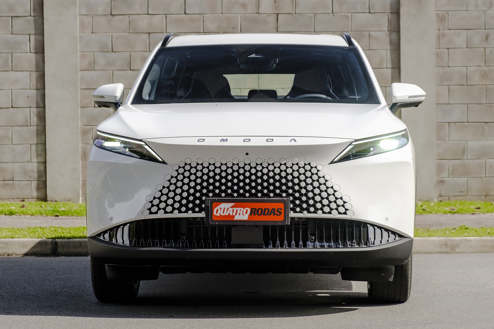 Carro branco Omoda de frente, com grade frontal em padr&atilde;o de colmeia e far&oacute;is finos acesos, estacionado em rua asfaltada com parede de tijolos ao fundo. Placa QUATRO RODAS no centro da grade.