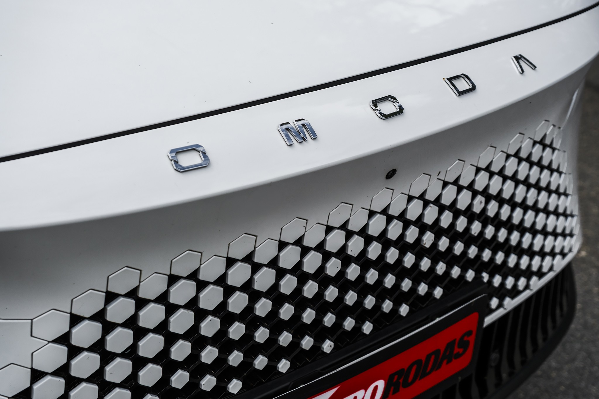 Traseira de carro branco com o nome OMODA em letras cromadas e uma grade inferior preta com padr&atilde;o de hex&aacute;gonos brancos e cinzas. Uma faixa vermelha com RODAS em branco aparece na parte inferior