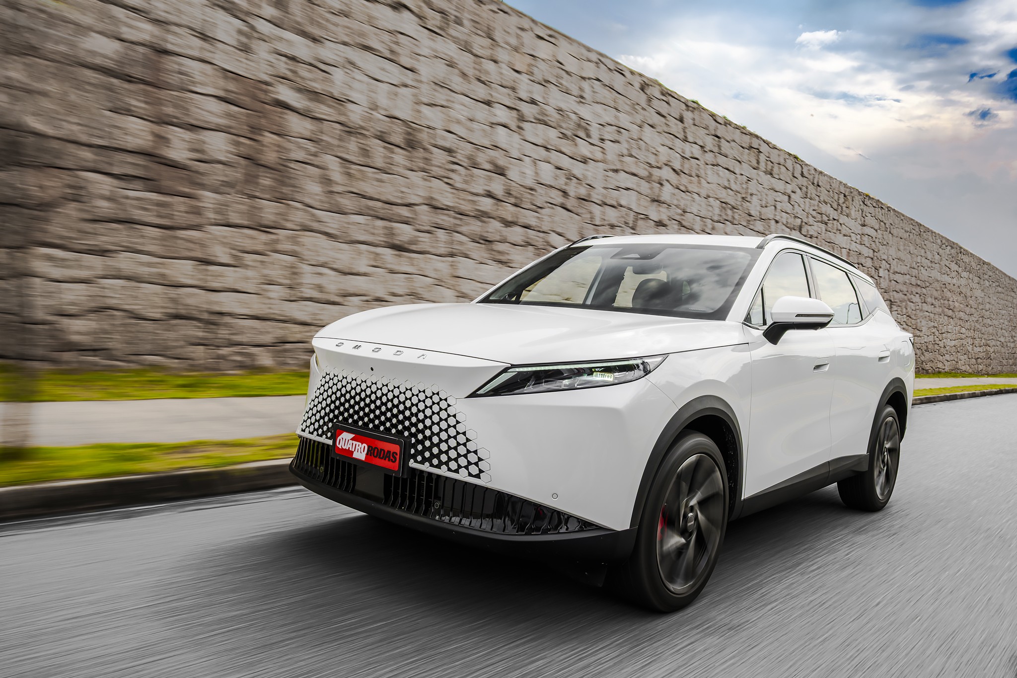 SUV branco moderno em movimento, visto de frente e lateral, com grade frontal em padr&atilde;o hexagonal e placa vermelha Auto Esporte. Ao fundo, um muro de pedras e c&eacute;u nublado, com asfalto borrado indicando velocidade