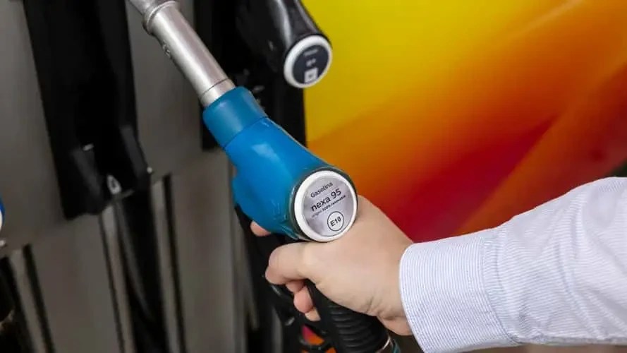 Mão de uma pessoa segurando um bico de bomba de combustível azul, com a etiqueta Gasolina nexa 95 E10 visível. Ao fundo, um painel de bomba de gasolina com detalhes em laranja e vermelho