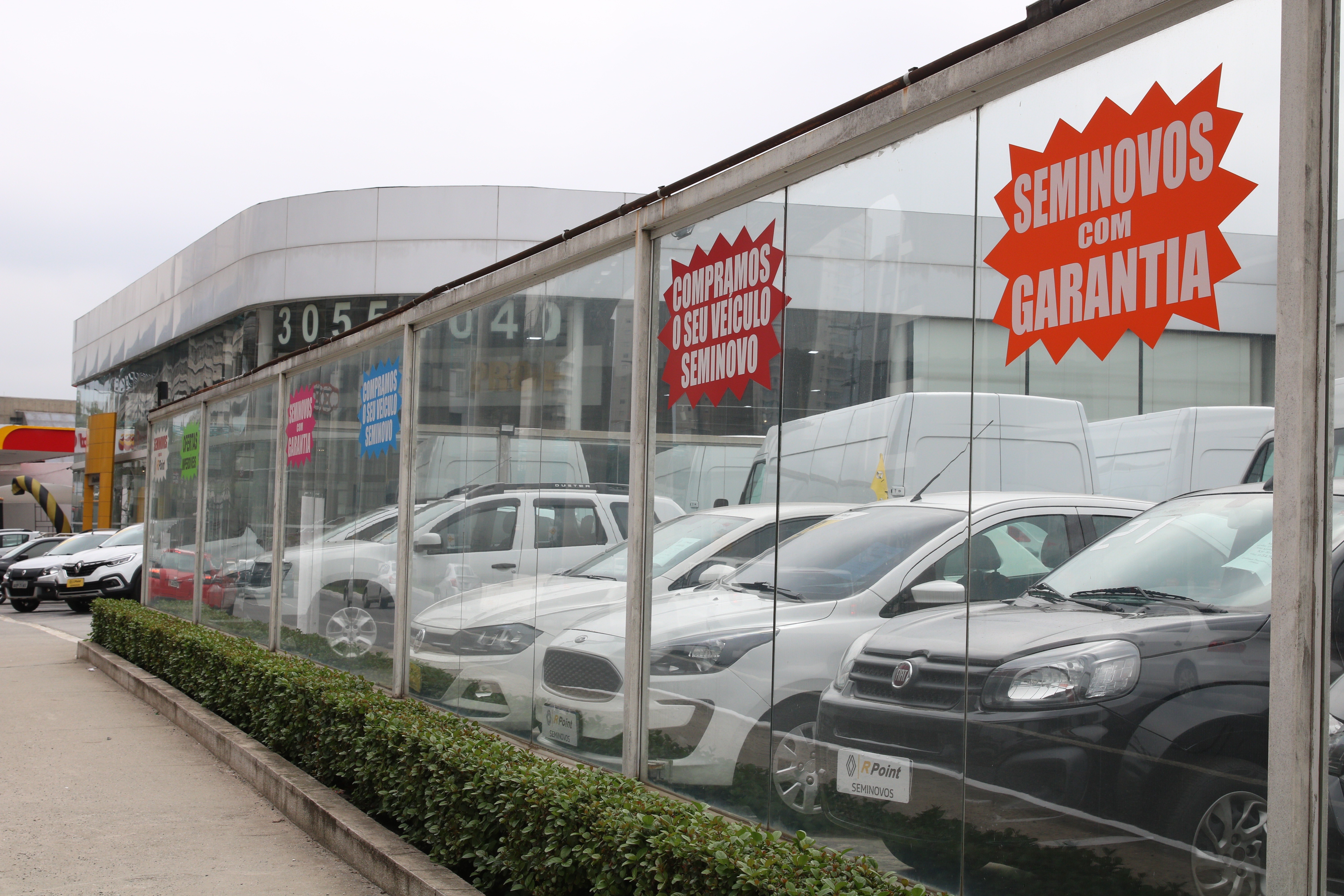 Fachada de concession&aacute;ria com carros seminovos expostos atr&aacute;s de vidros. Adesivos vermelhos em formato de estrela anunciam COMPRAMOS O SEU VE&Iacute;CULO SEMINOVO e SEMINOVOS COM GARANTIA. Uma fileira de arbustos verdes decora a frente do estabelecimento.