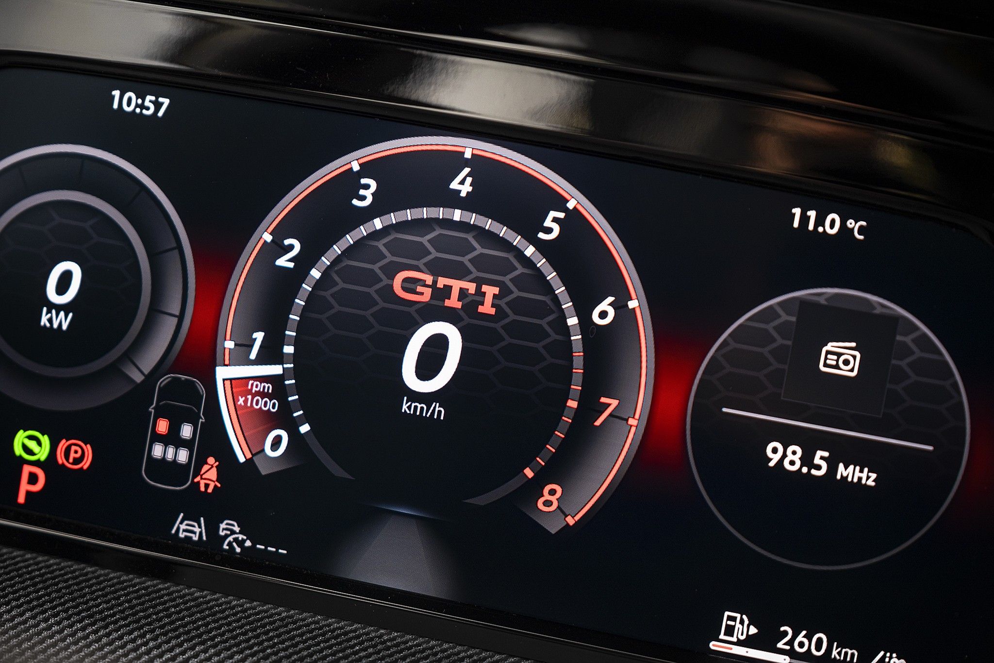 Painel digital de carro com veloc&iacute;metro central marcando 0 km/h e logo GTI em laranja. &Agrave; esquerda, medidor de pot&ecirc;ncia em 0 kW. &Agrave; direita, r&aacute;dio em 98.5 MHz e temperatura externa de 11.0&deg;C. Luzes de advert&ecirc;ncia e autonomia de 260 km vis&iacute;veis na parte inferior