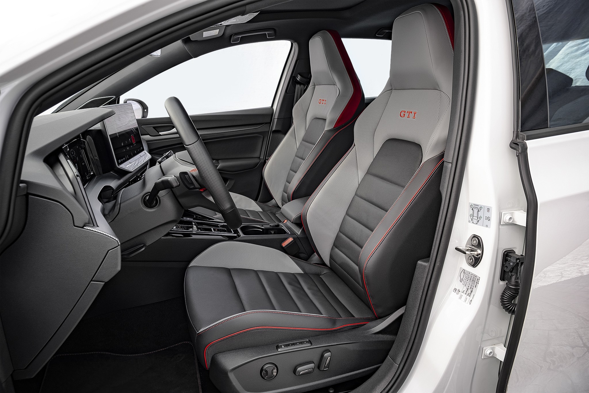 Interior de um carro branco, mostrando o banco do motorista e do passageiro em cinza e preto com costuras vermelhas, e o volante preto. O painel possui uma tela digital e o teto solar est&aacute; vis&iacute;vel