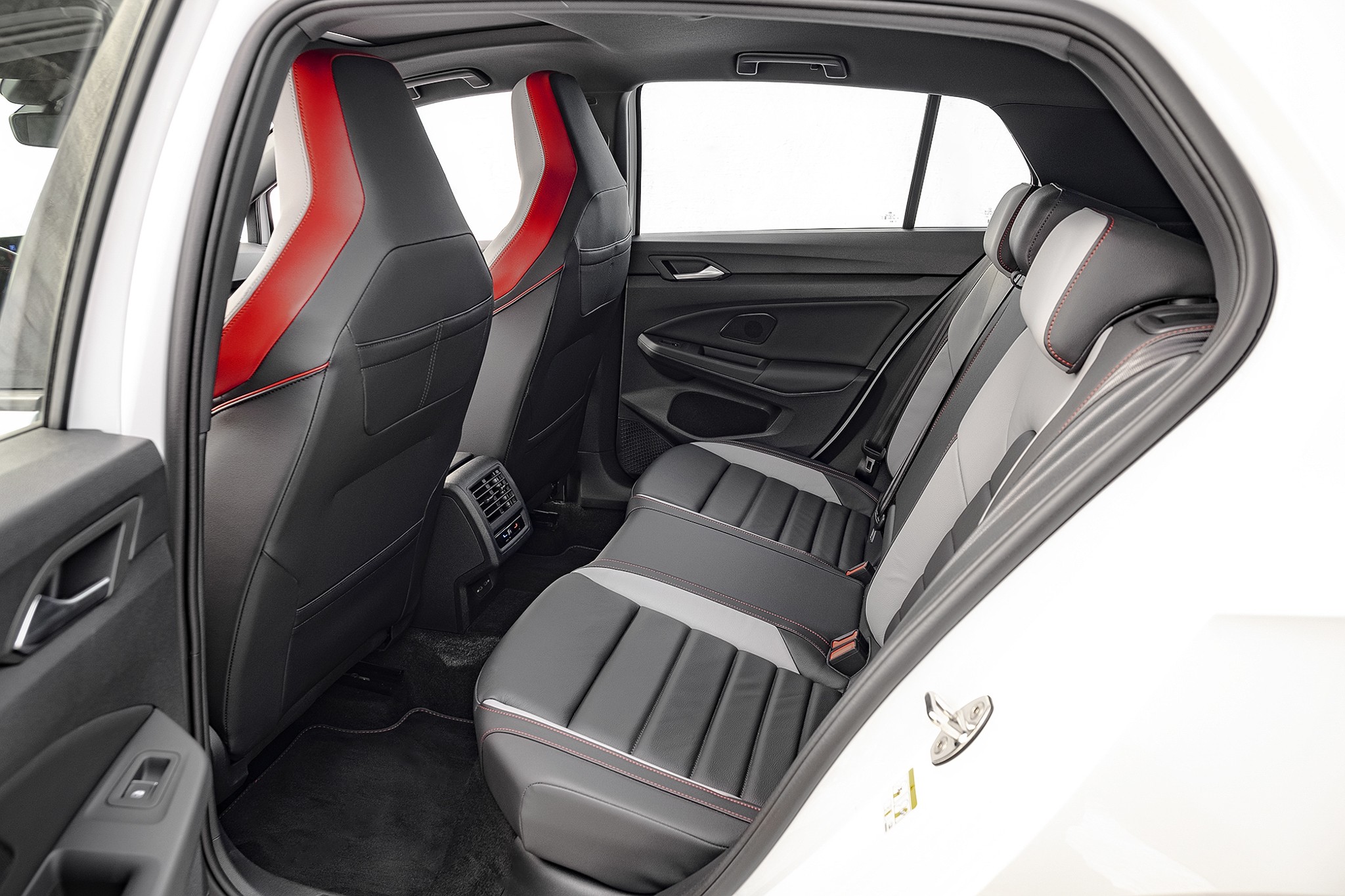 Interior de um carro branco, mostrando os bancos traseiros e parte dos dianteiros. Os bancos s&atilde;o pretos com detalhes em cinza e costuras vermelhas, e os encostos dos bancos dianteiros t&ecirc;m uma faixa vermelha vertical. O assoalho &eacute; preto, e as janelas mostram um fundo branco