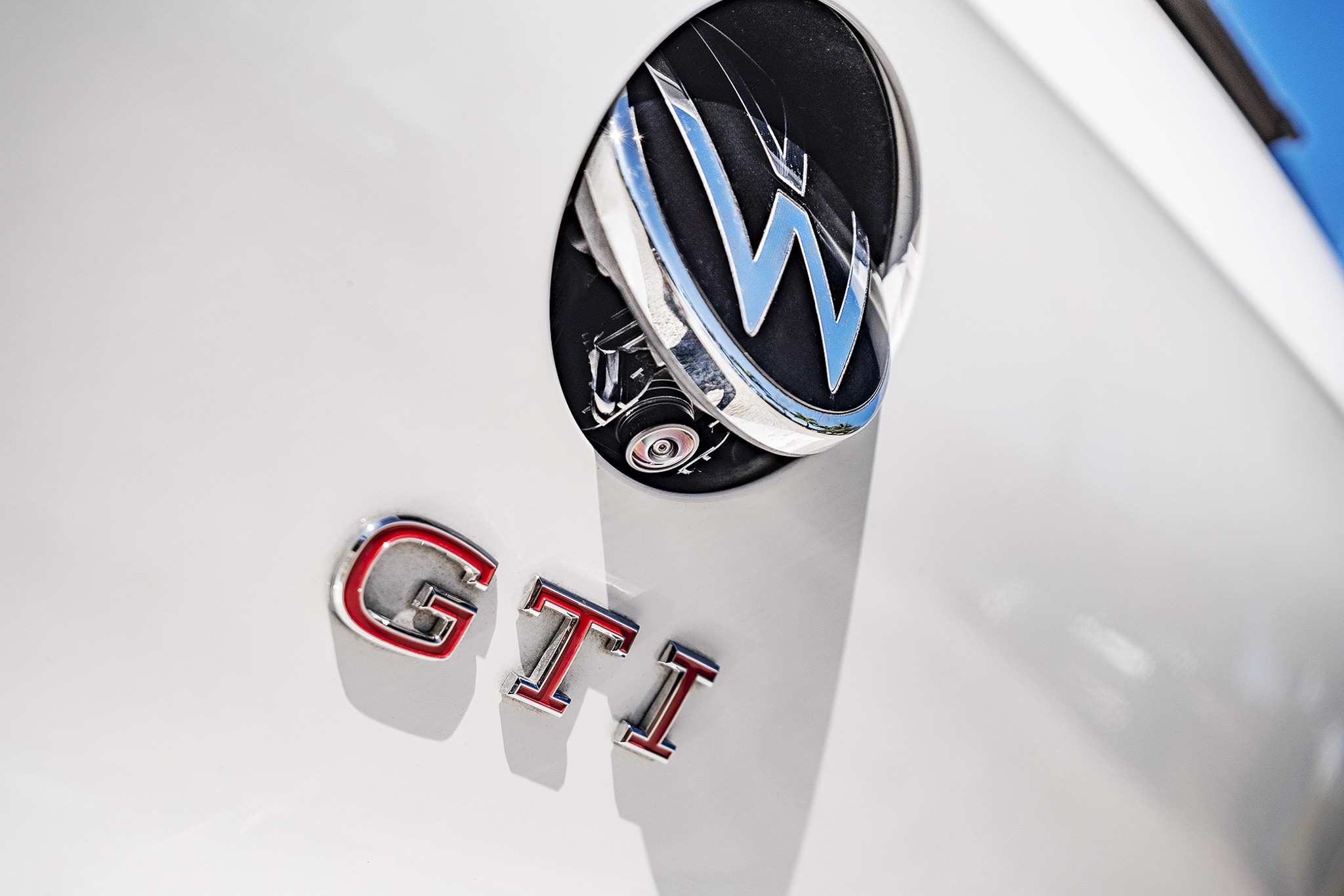 Emblema redondo da Volkswagen com o logo VW em azul e prata, parcialmente aberto, revelando uma c&acirc;mera interna. Abaixo, as letras GTI em vermelho e prata, sobre uma superf&iacute;cie branca brilhante