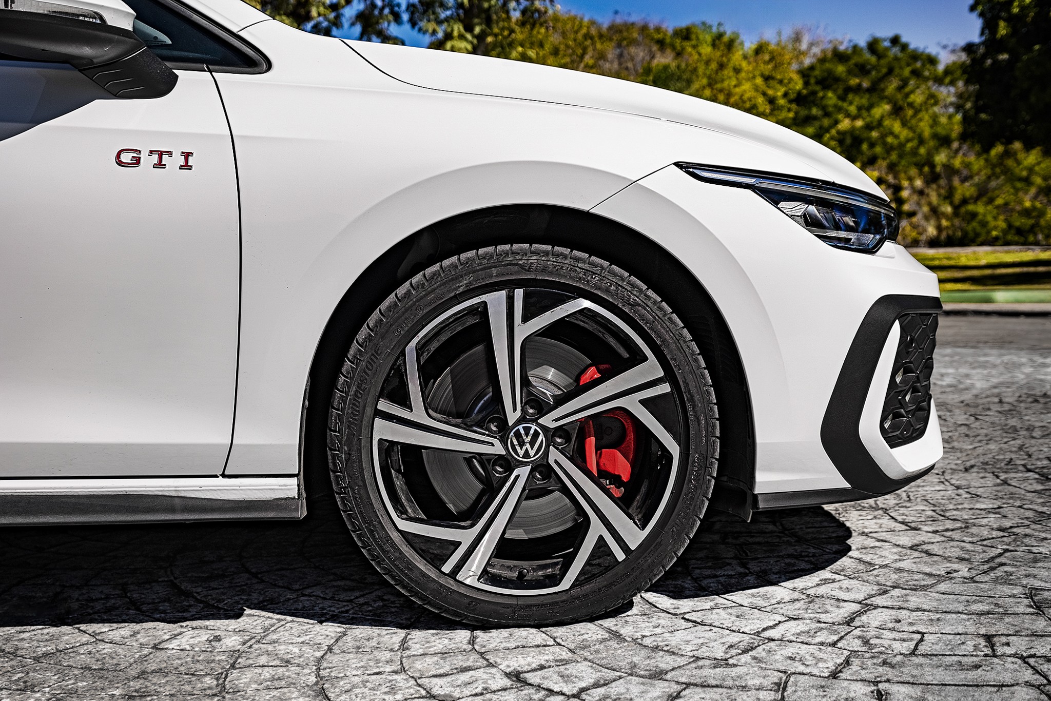 Lateral dianteira de um carro branco com emblema GTI em vermelho, roda preta e prata com pin&ccedil;a de freio vermelha, farol afilado e grade frontal preta, estacionado em piso de paralelep&iacute;pedos sob luz solar