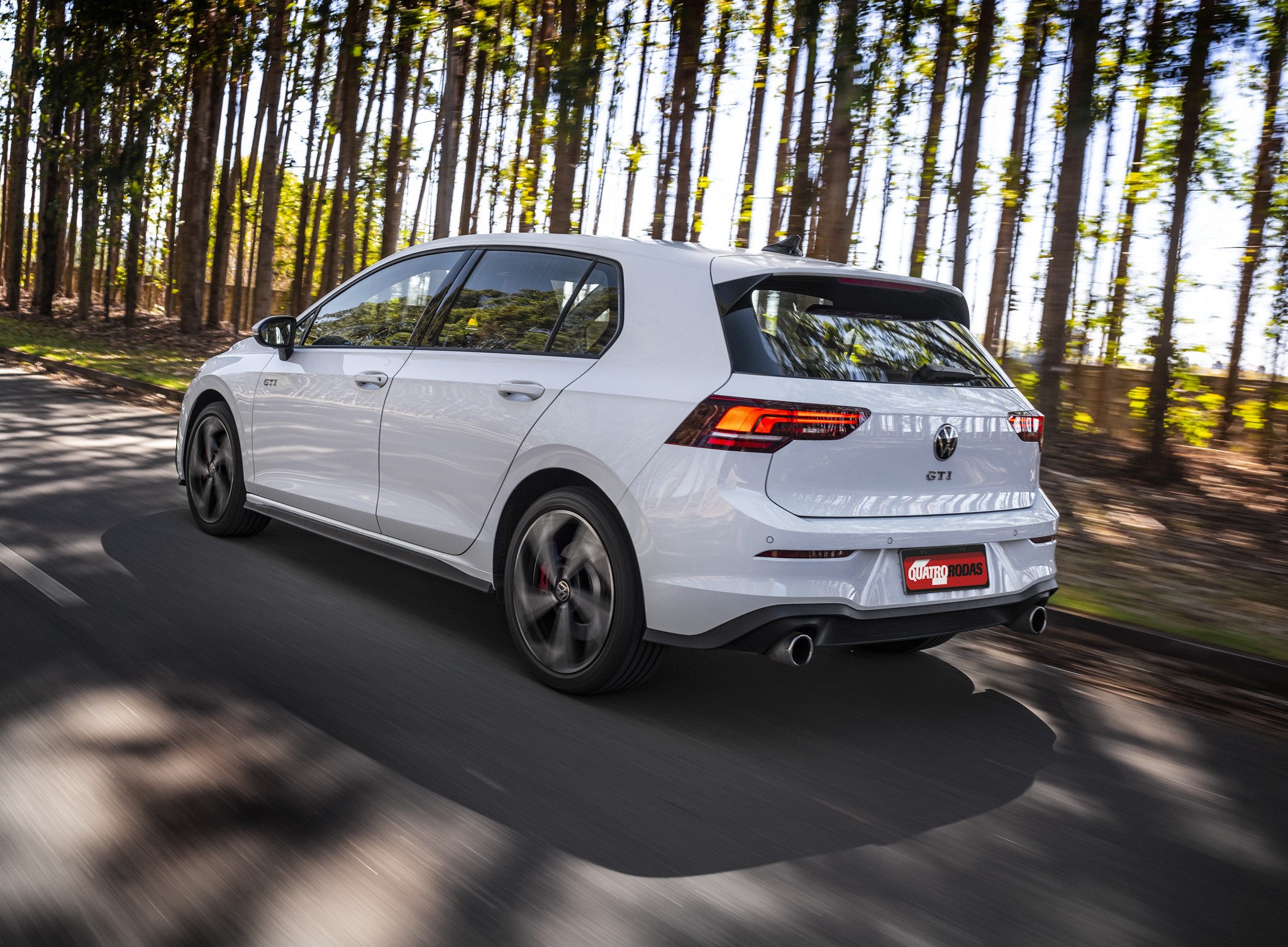 Carro hatch branco Volkswagen Golf GTI em movimento, visto de traseira lateral, em uma estrada asfaltada com &aacute;rvores altas e finas ao fundo, sob luz solar