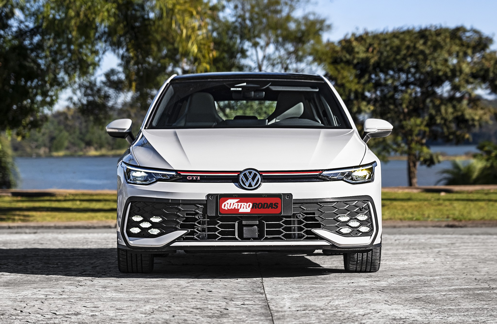Volkswagen Golf GTI branco, visto de frente, com grade preta em colmeia e faixa vermelha. O carro est&aacute; estacionado em um piso de concreto, com &aacute;rvores e um lago ao fundo