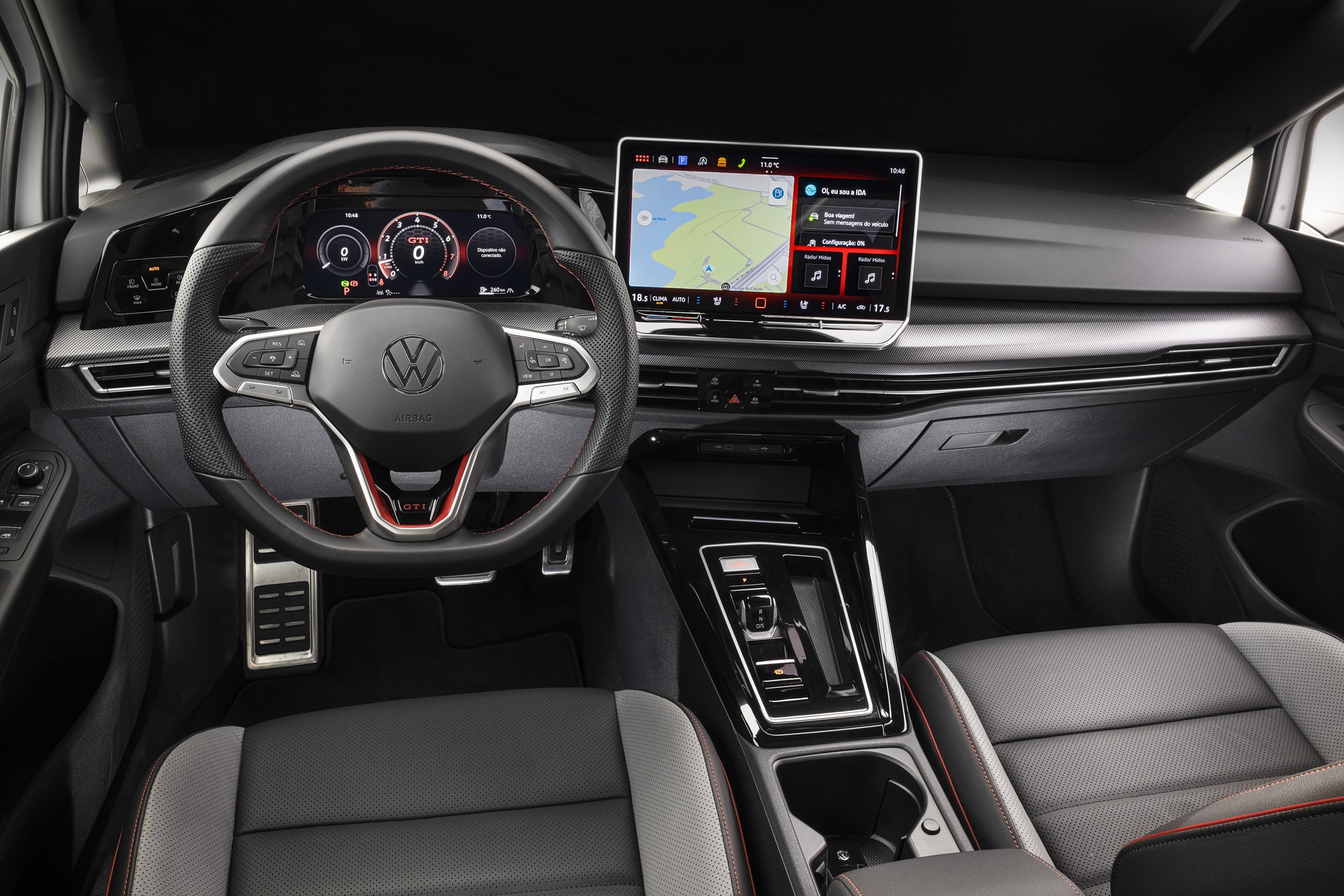 Interior de um carro moderno, com volante multifuncional preto e detalhes vermelhos, painel digital exibindo veloc&iacute;metro e conta-giros, e uma tela multim&iacute;dia flutuante com mapa de navega&ccedil;&atilde;o e controles de &aacute;udio. O console central tem acabamento em preto brilhante e o banco do motorista &eacute; cinza e preto com costura vermelha