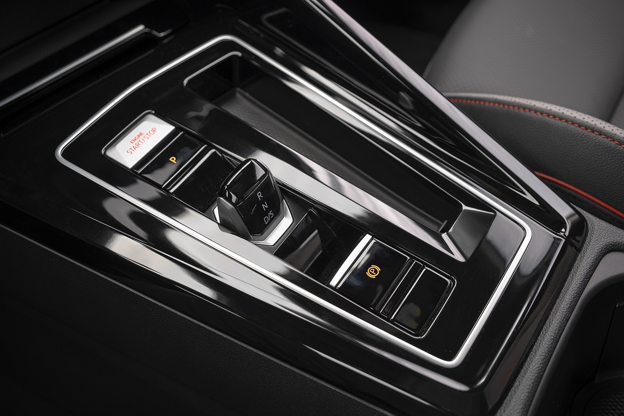 Console central de um carro com acabamento preto brilhante, destacando o seletor de marchas compacto, bot&otilde;es de partida e freio de estacionamento eletr&ocirc;nico, e um banco de couro escuro com costura vermelha ao fundo