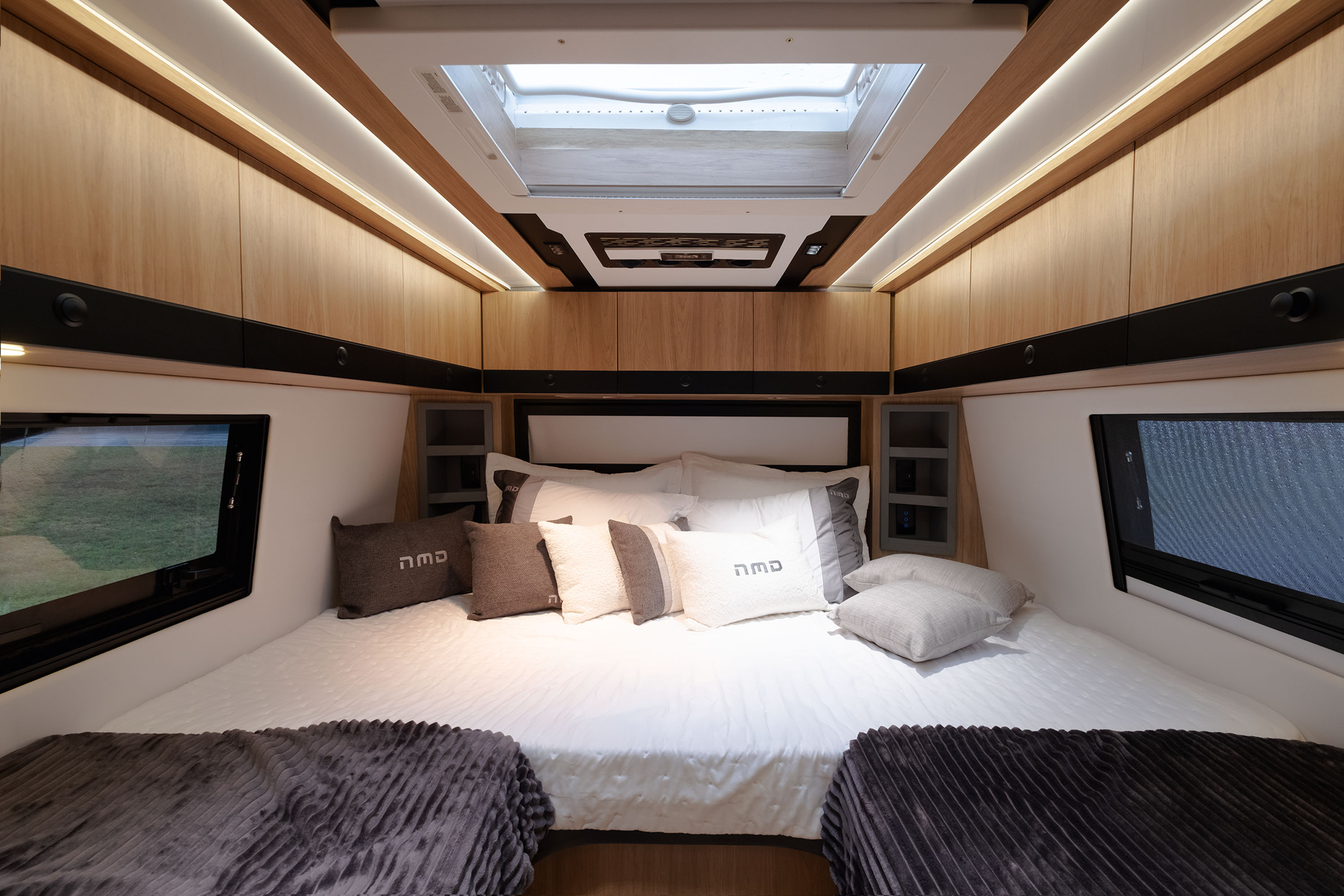 Interior de um quarto de motorhome com cama de casal central, len&ccedil;&oacute;is brancos, travesseiros cinzas e brancos com a inscri&ccedil;&atilde;o nmd, e cobertores cinza escuro nas laterais. Arm&aacute;rios de madeira clara com ilumina&ccedil;&atilde;o embutida e uma claraboia no teto. Janelas retangulares escuras nas laterais mostram a paisagem verde externa