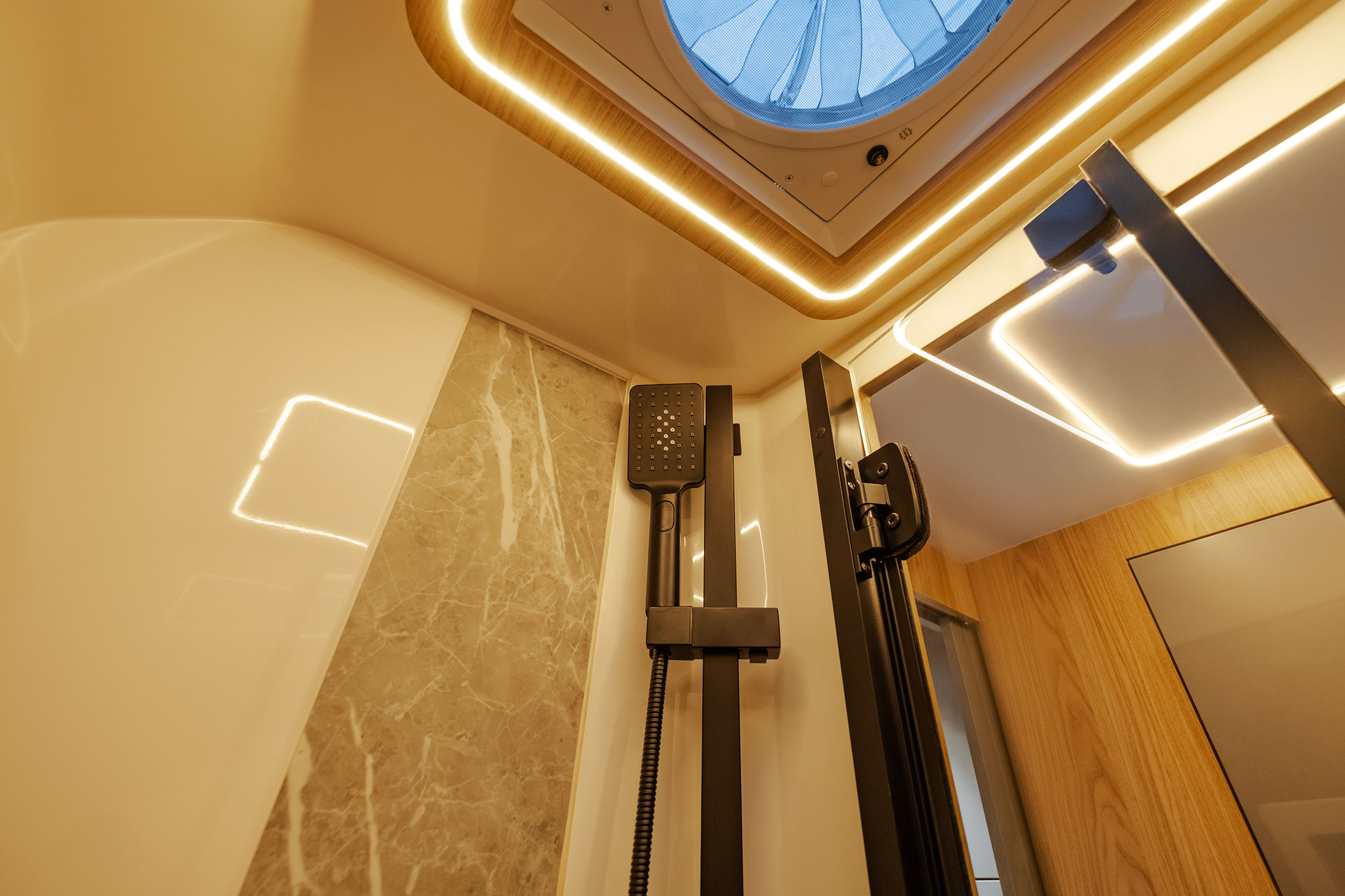 Interior de um banheiro moderno com chuveiro preto fosco, paredes claras e detalhes em m&aacute;rmore. O teto possui uma claraboia redonda com ventilador e ilumina&ccedil;&atilde;o LED embutida, criando um ambiente sofisticado e funcional