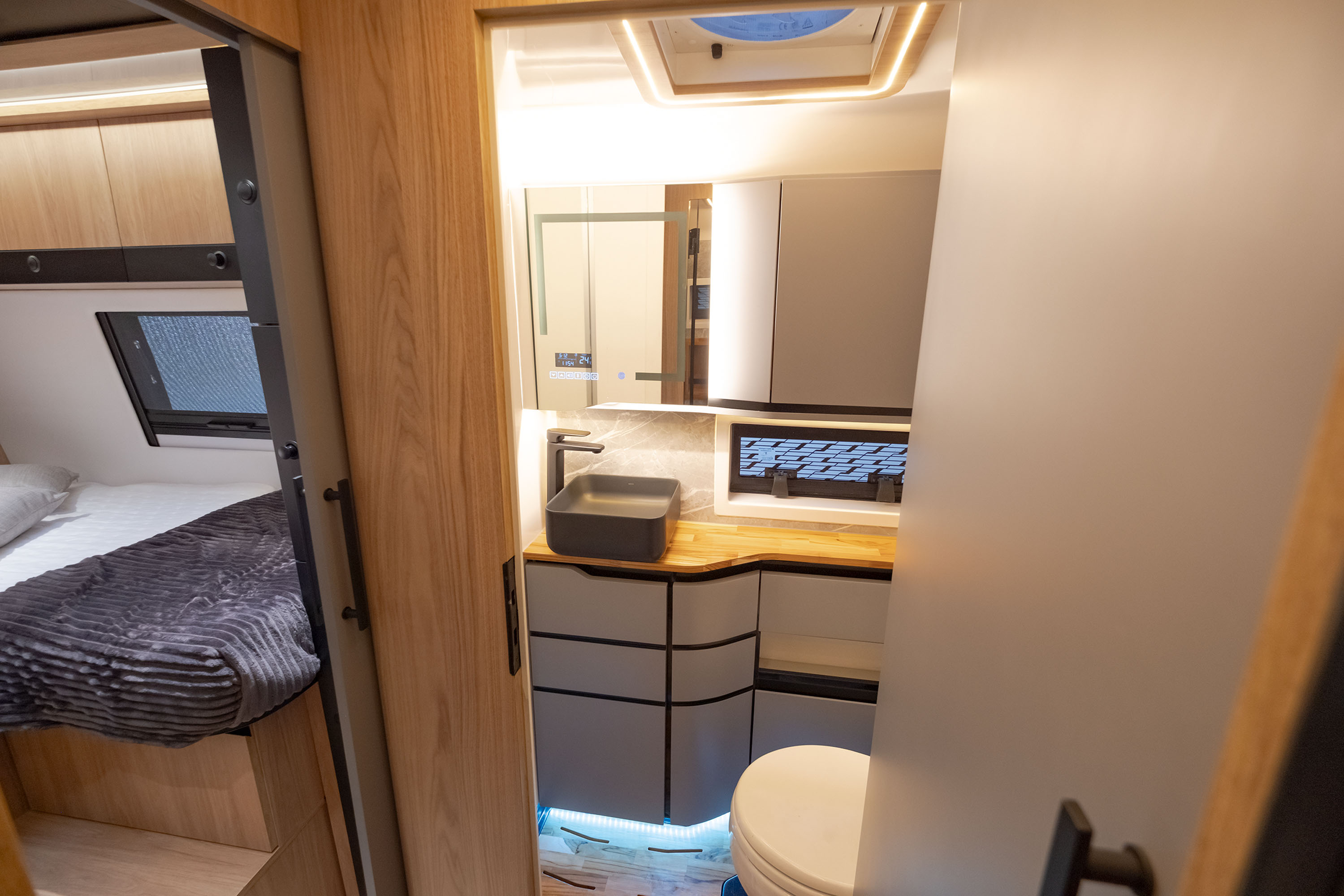 Interior de um motorhome moderno, mostrando um quarto com cama e um banheiro compacto. O quarto tem uma cama com len&ccedil;&oacute;is brancos e uma manta cinza escura, com arm&aacute;rios de madeira clara acima. O banheiro, &agrave; direita, possui uma pia retangular cinza escura sobre um balc&atilde;o de madeira, arm&aacute;rios cinzas e um vaso sanit&aacute;rio branco. H&aacute; ilumina&ccedil;&atilde;o embutida no teto e sob os arm&aacute;rios do banheiro, criando um ambiente acolhedor