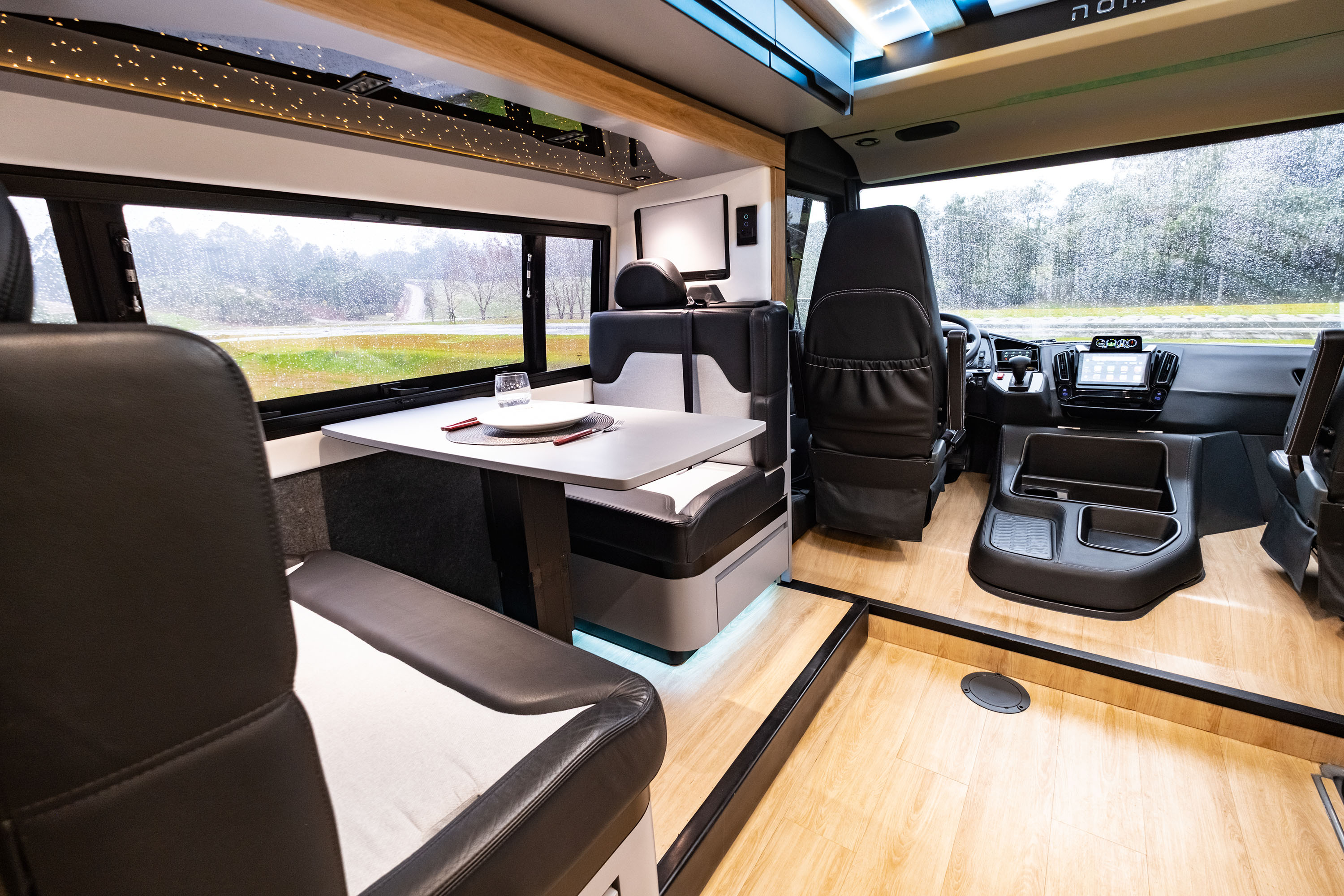 Interior de um motorhome moderno com piso de madeira clara, bancos estofados em preto e branco, e uma mesa com prato e copo. Ao fundo, janelas mostram uma paisagem verde com &aacute;rvores e uma estrada, e o painel de controle do ve&iacute;culo.