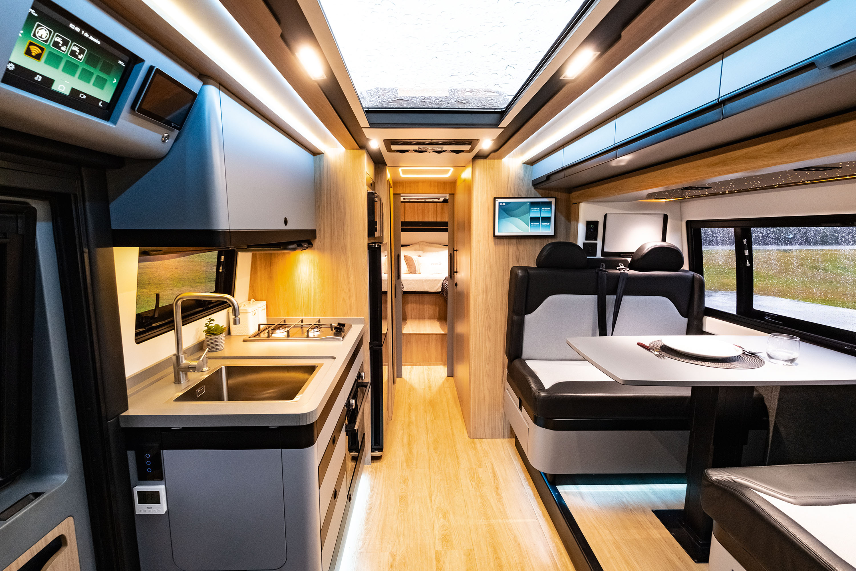 Interior moderno de um motorhome com cozinha compacta &agrave; esquerda, incluindo pia e fog&atilde;o. &Agrave; direita, assentos pretos e brancos com uma mesa posta. Ao fundo, um corredor leva a uma &aacute;rea de dormir com beliches. O ambiente &eacute; iluminado por luzes LED no teto e sob os m&oacute;veis, e h&aacute; telas digitais exibindo informa&ccedil;&otilde;es