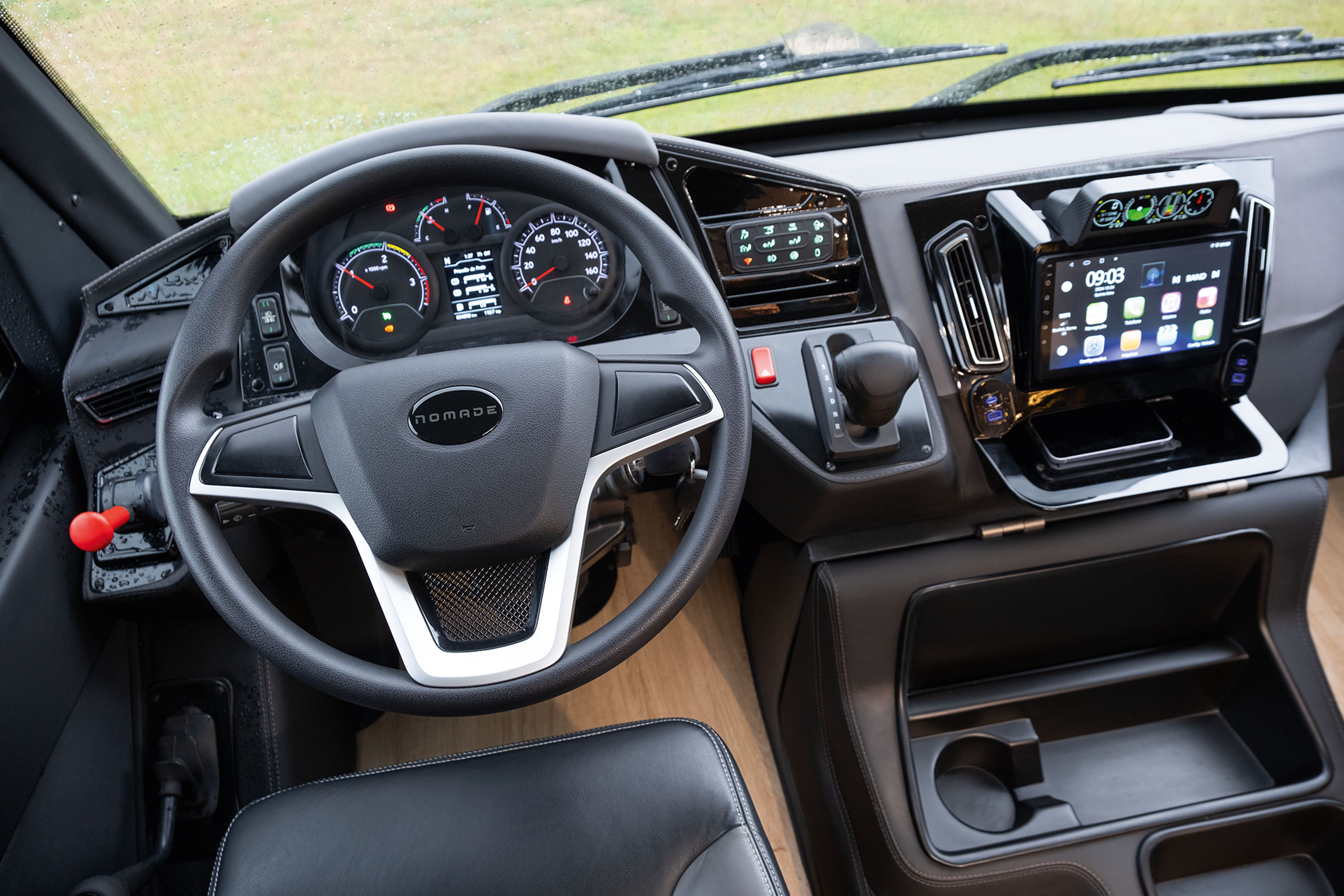 Interior de um ve&iacute;culo com volante preto e detalhes prateados, painel de instrumentos digital, tela multim&iacute;dia com &iacute;cones de aplicativos e um smartphone em um compartimento abaixo. &Agrave; direita, c&acirc;mbio autom&aacute;tico e porta-copos.