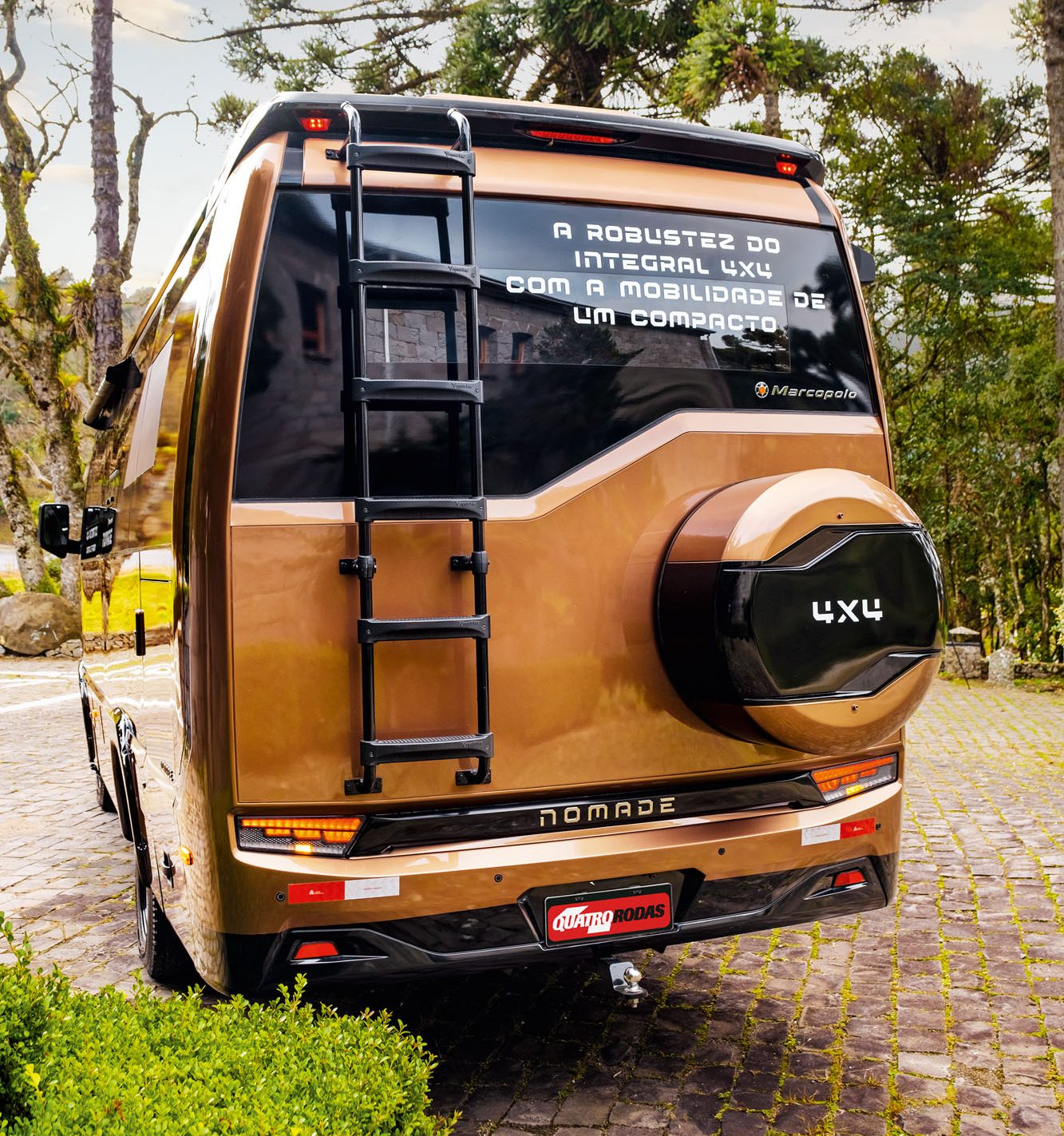 Traseira de um motorhome Marcopolo Nomade 4x4 cor bronze, com escada preta, estepe e a frase A robustez do integral 4x4 com a mobilidade de um compacto no vidro. Estacionado em p&aacute;tio de pedras, com arbustos e &aacute;rvores altas ao redor