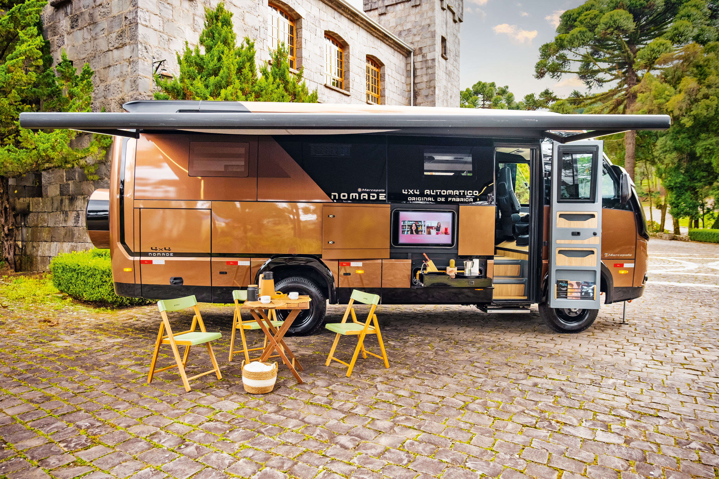 Motorhome Nomade marrom e preto estacionado em p&aacute;tio de pedras, com toldo aberto e mesa de madeira com cadeiras dobr&aacute;veis &agrave; frente. A lateral do ve&iacute;culo exibe uma TV e compartimentos abertos, revelando utens&iacute;lios de cozinha. Ao fundo, um edif&iacute;cio de pedra com janelas e &aacute;rvores verdes