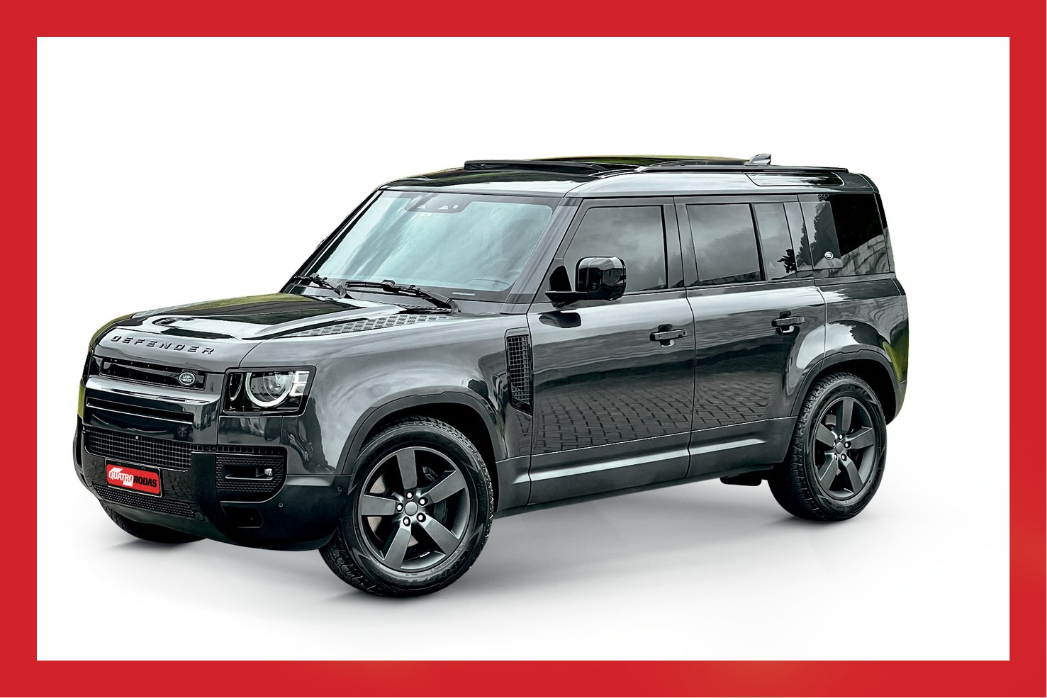 SUV Land Rover Defender cinza-escuro, com teto solar e rodas pretas, estacionado em piso de paralelep&iacute;pedos, visto de perfil em &acirc;ngulo frontal, contra fundo branco
