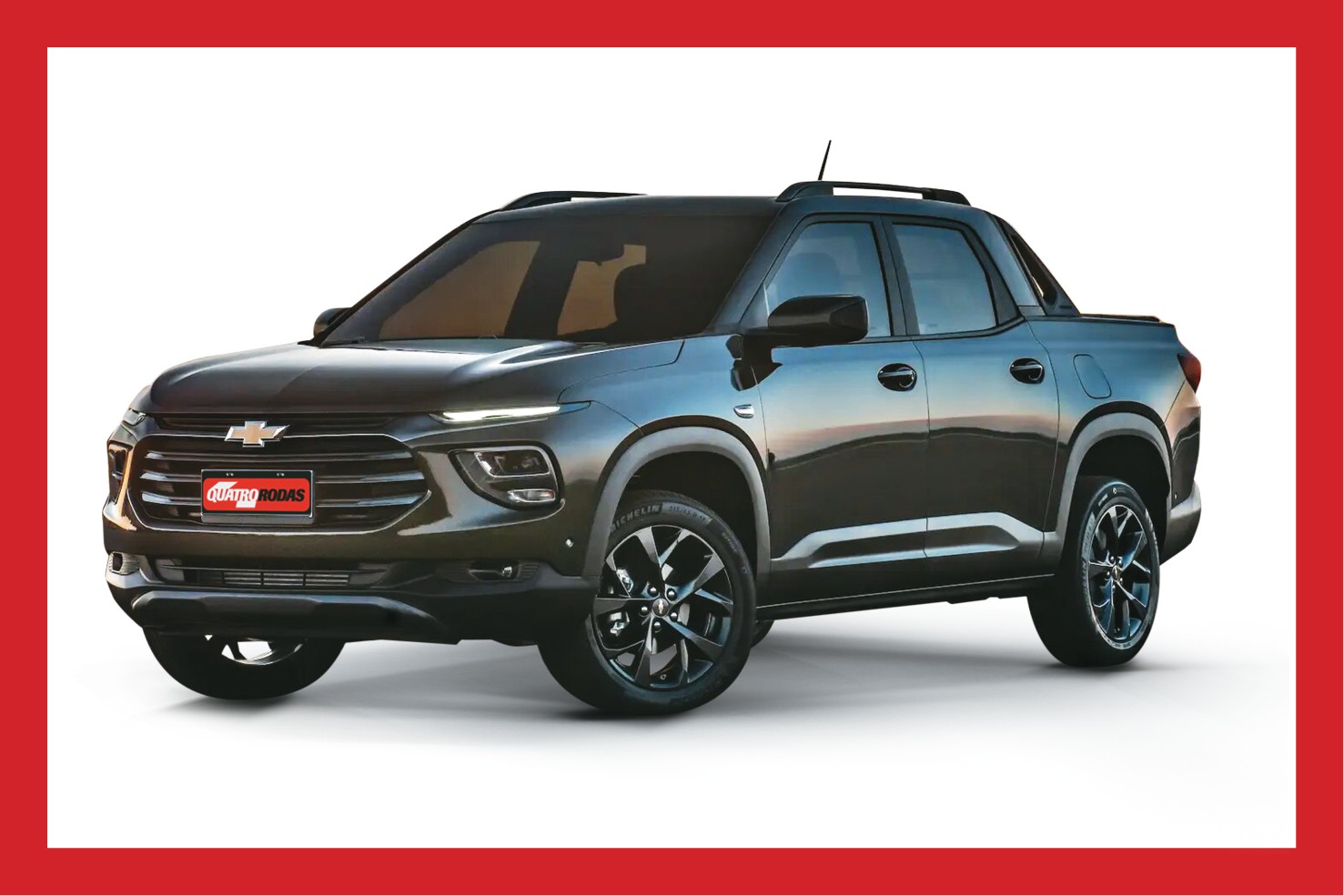 Picape Chevrolet Montana cinza escura, vista de tr&ecirc;s quartos frontal, com far&oacute;is acesos, grade preta e placa vermelha Quatro Rodas na frente. Possui rack de teto e rodas esportivas pretas, destacando-se em fundo branco