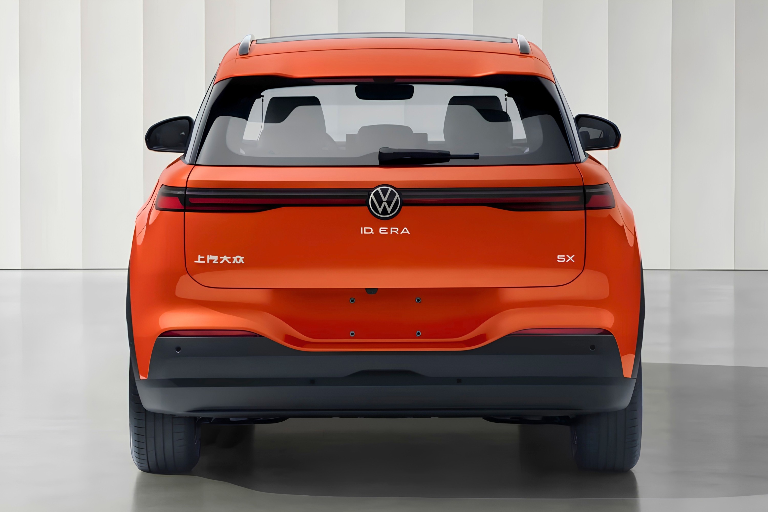 Traseira de um SUV el&eacute;trico Volkswagen ID.ERA laranja, com o logo VW e o nome do modelo centralizados. &Agrave; esquerda, caracteres chineses e &agrave; direita, 5X. O carro est&aacute; em um est&uacute;dio com paredes brancas e piso cinza