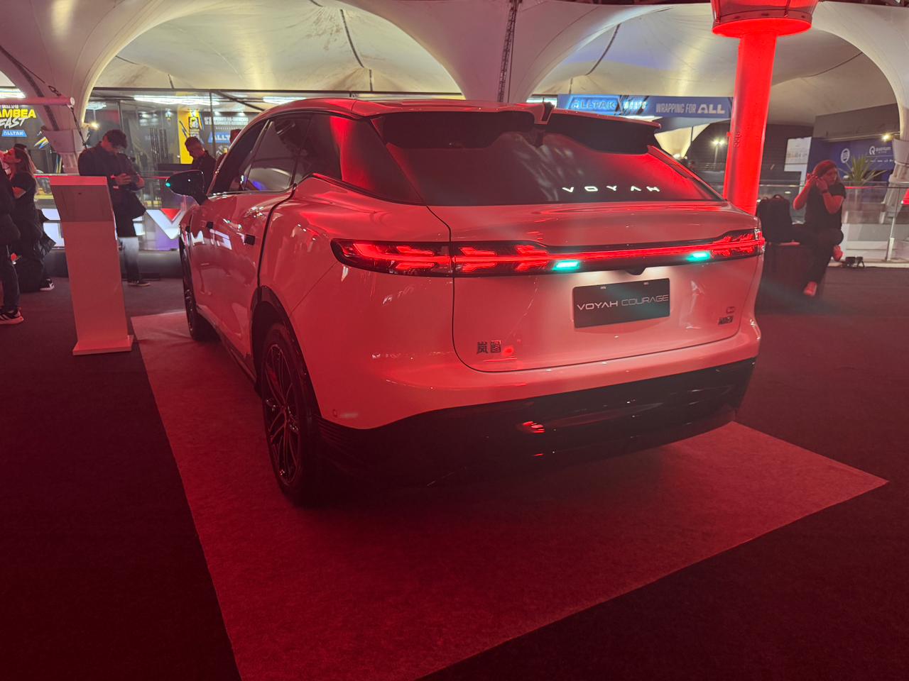 Traseira de um carro SUV branco, modelo Voyah Free, com lanternas vermelhas e uma faixa de luz azul. O carro est&aacute; em um evento noturno, sob uma tenda branca, com ilumina&ccedil;&atilde;o vermelha no ch&atilde;o e pessoas ao fundo.