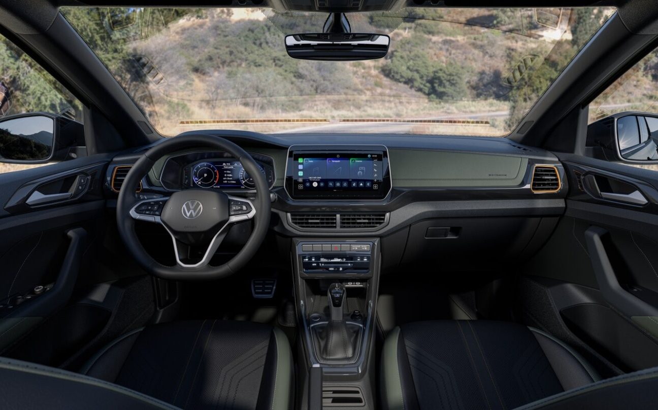 Vis&atilde;o interna do painel de um carro Volkswagen, com volante multifuncional, painel digital, central multim&iacute;dia e c&acirc;mbio autom&aacute;tico. O interior &eacute; predominantemente preto com detalhes em cinza e dourado, e o para-brisa mostra uma estrada com vegeta&ccedil;&atilde;o ao fundo