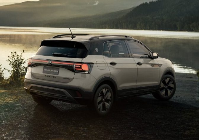 SUV branco estacionado em uma estrada de terra &agrave; beira de um lago, com montanhas e &aacute;rvores ao fundo. O sol se p&otilde;e no horizonte, refletindo na &aacute;gua e criando um brilho dourado. O c&eacute;u tem nuvens escuras e claras, e a atmosfera &eacute; serena