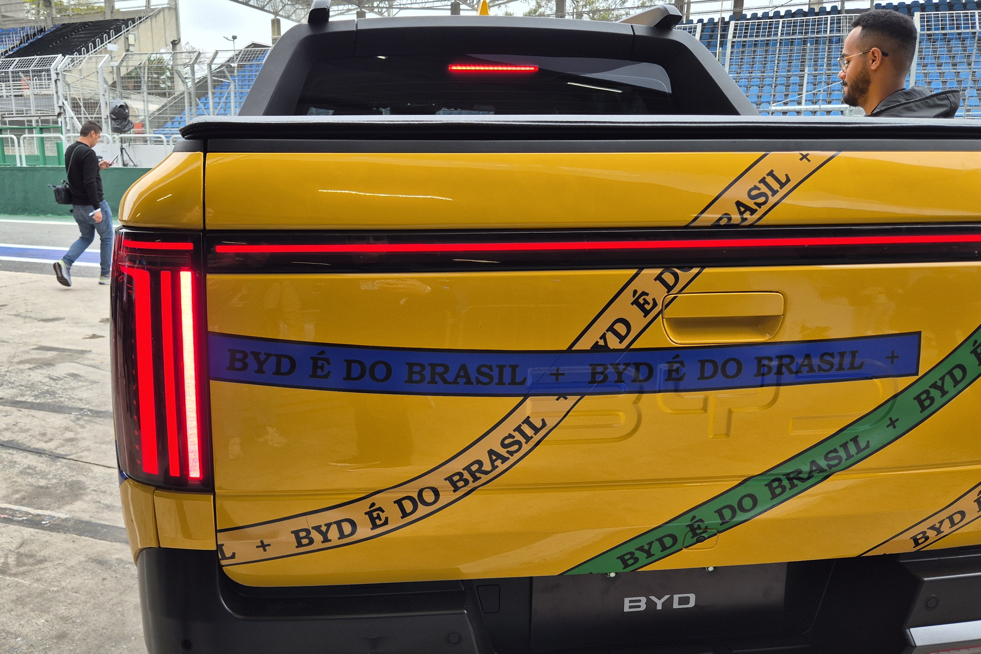 Traseira de uma picape amarela com adesivos BYD &Eacute; DO BRASIL em faixas azuis, verdes e pretas. Um homem com barba e &oacute;culos est&aacute; no banco do passageiro, e outro homem caminha ao fundo, pr&oacute;ximo a uma arquibancada
