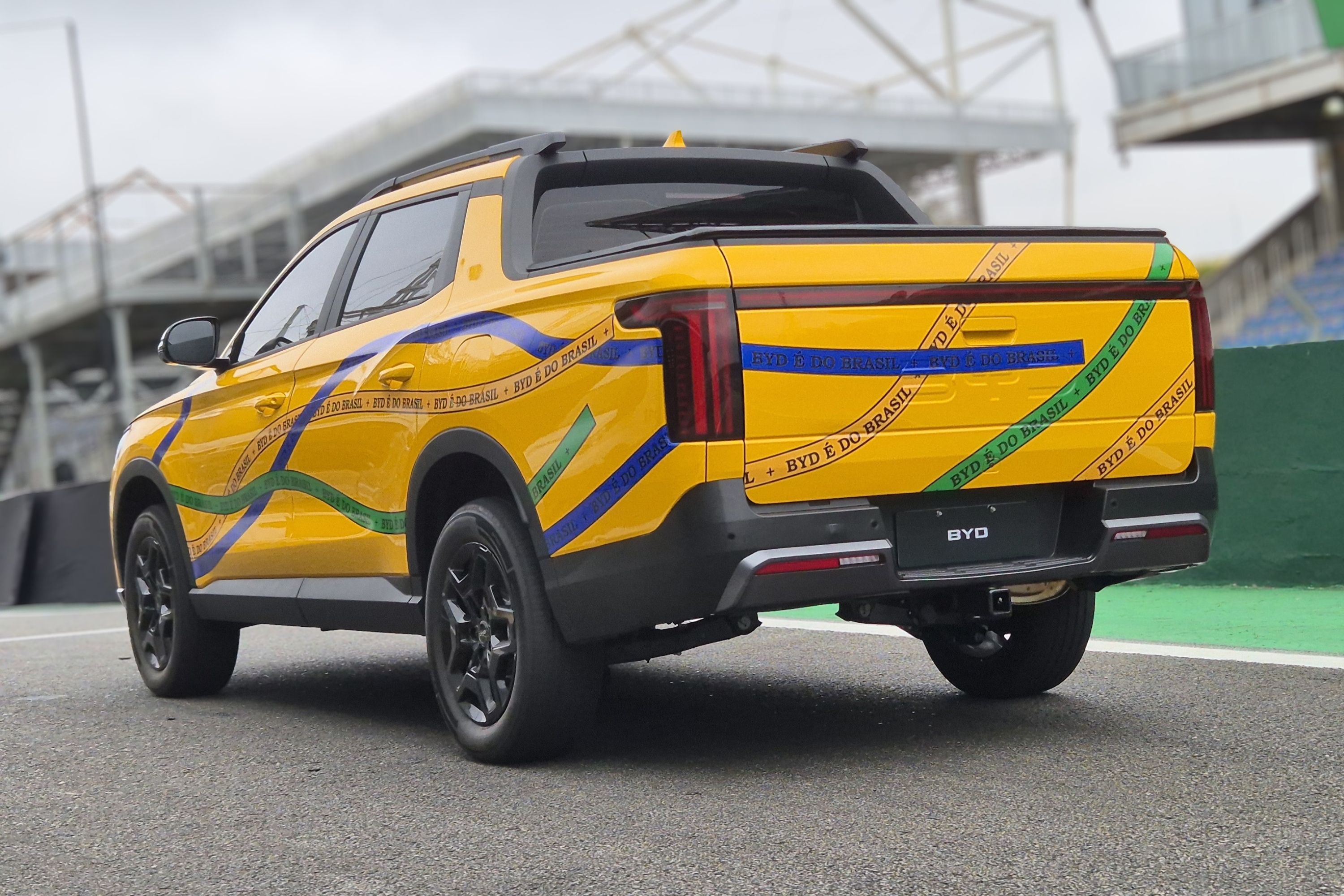 Picape amarela com faixas azuis e verdes, simulando a bandeira do Brasil, e a frase BYD &Eacute; DO BRASIL repetida, estacionada em uma pista de corrida com arquibancadas ao fundo