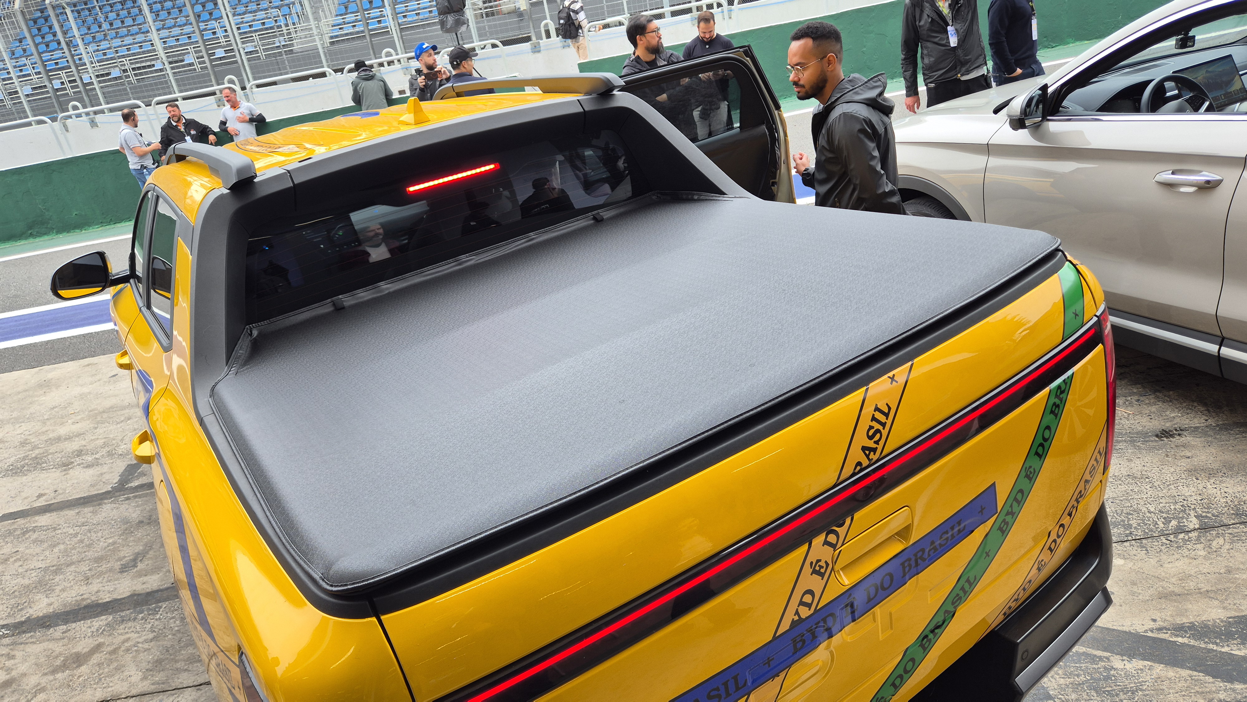 Picape amarela com capota mar&iacute;tima preta, adesivos de fitas do Senhor do Bonfim nas cores da bandeira do Brasil na traseira, em uma pista de corrida. Um homem de &oacute;culos e jaqueta preta est&aacute; ao lado da porta aberta do ve&iacute;culo, enquanto outras pessoas observam ao fundo