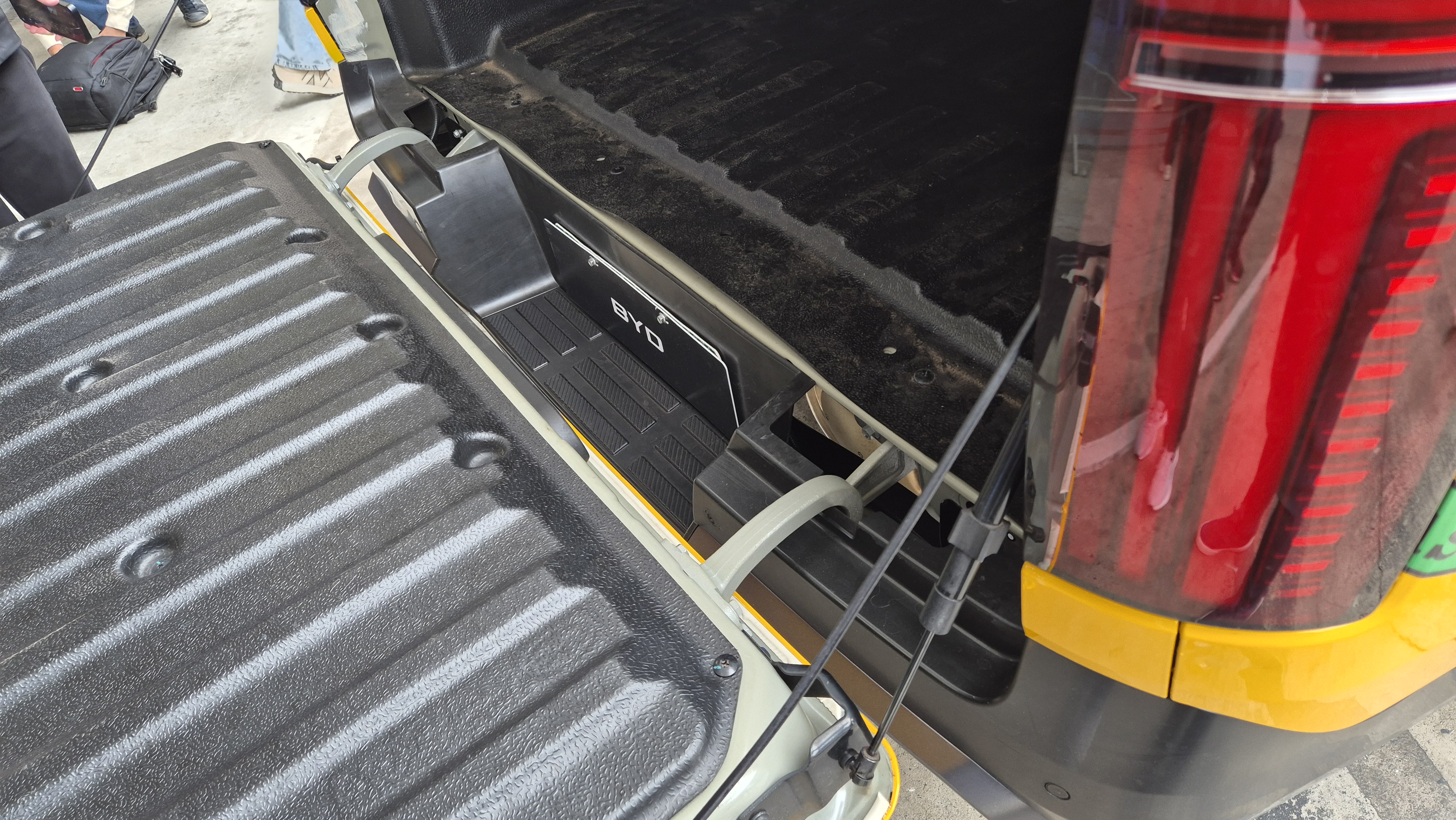Ca&ccedil;amba de picape com a tampa traseira aberta, revelando um compartimento lateral com o logo BYD e um piso texturizado. A lanterna traseira vermelha do ve&iacute;culo &eacute; vis&iacute;vel &agrave; direita, com reflexos de pessoas.
