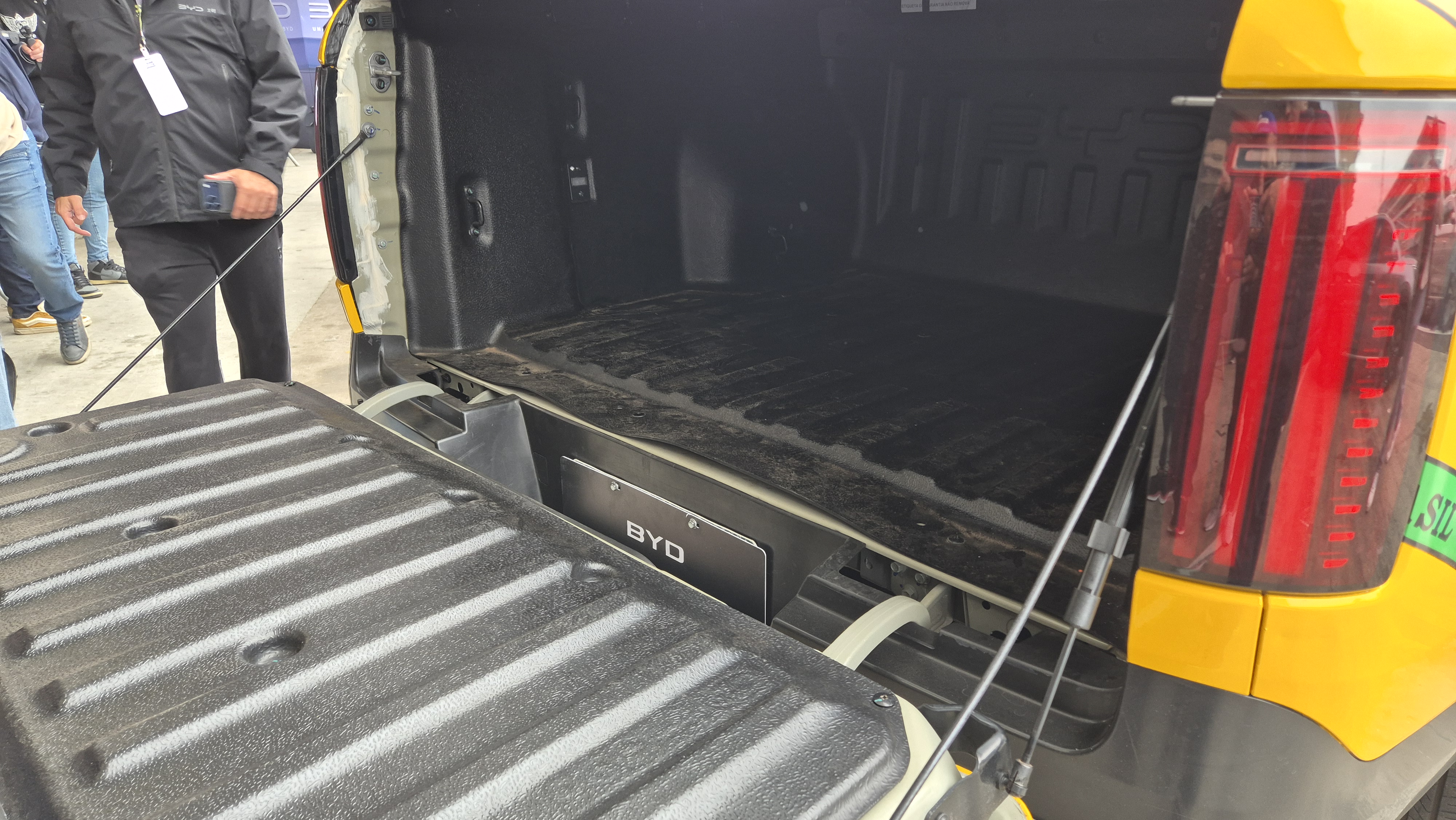 Ca&ccedil;amba de uma picape amarela com a tampa traseira aberta, revelando o interior escuro e texturizado. Uma placa preta com o nome BYD est&aacute; vis&iacute;vel na parte inferior da ca&ccedil;amba. Uma pessoa de jaqueta preta e cal&ccedil;a escura est&aacute; &agrave; esquerda, segurando um celular